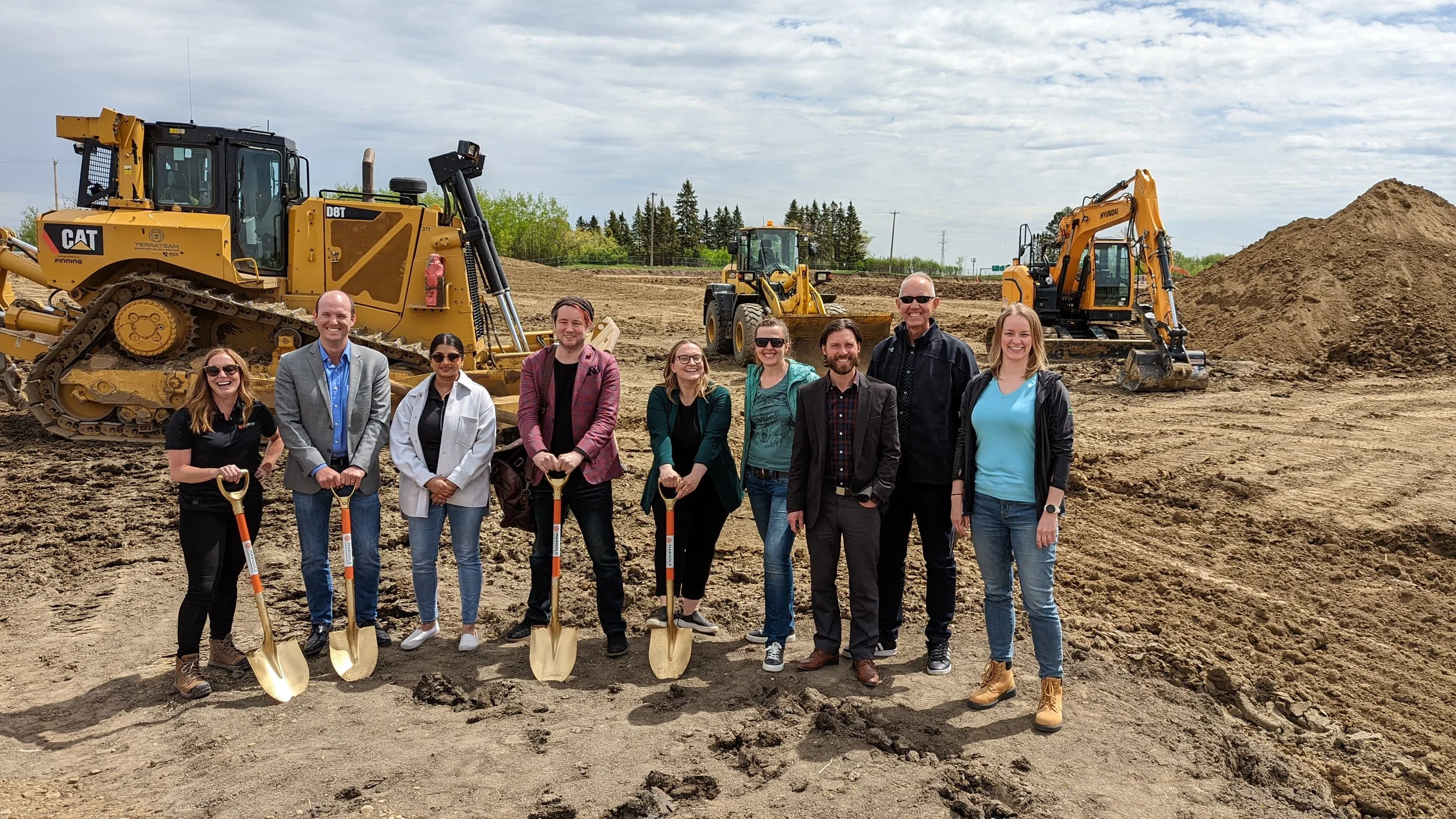 Group of ten people standing with shovels in front of construction equipment and dirt piles at a construction site, smiling.