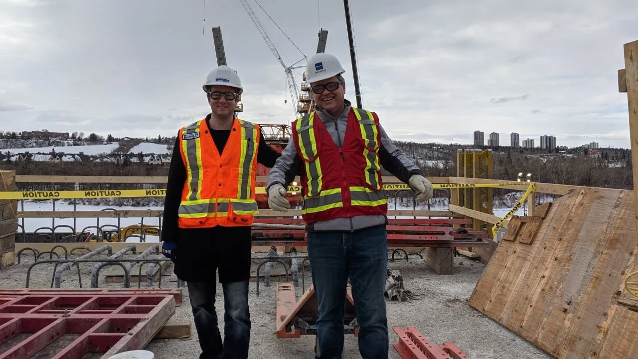 Two construction workers wearing hard hats and safety vests smiling on a construction site with cranes and a cityscape in the background.