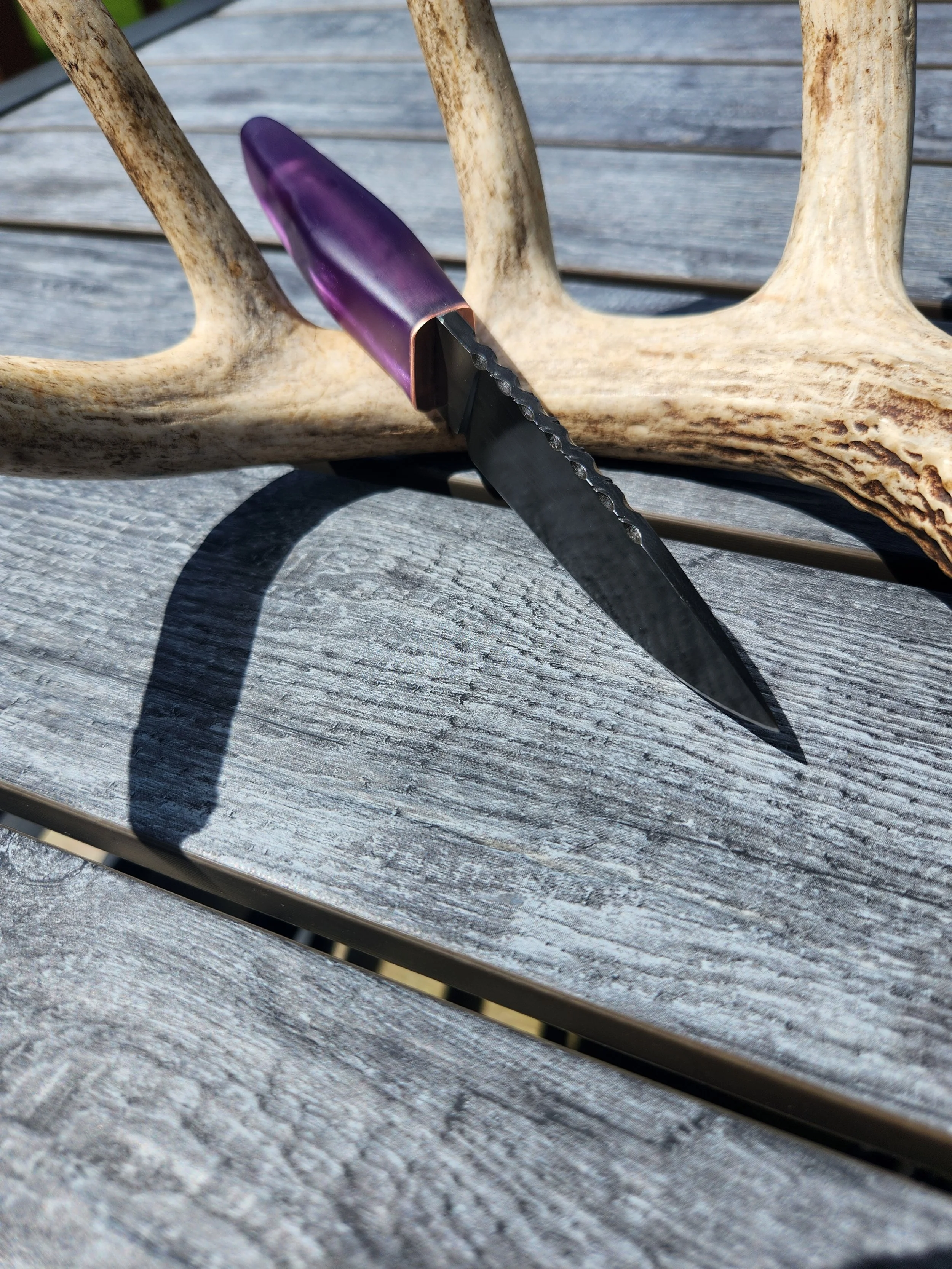 A small black knife with a purple handle resting on a weathered wooden surface, with a detached deer antler behind it.