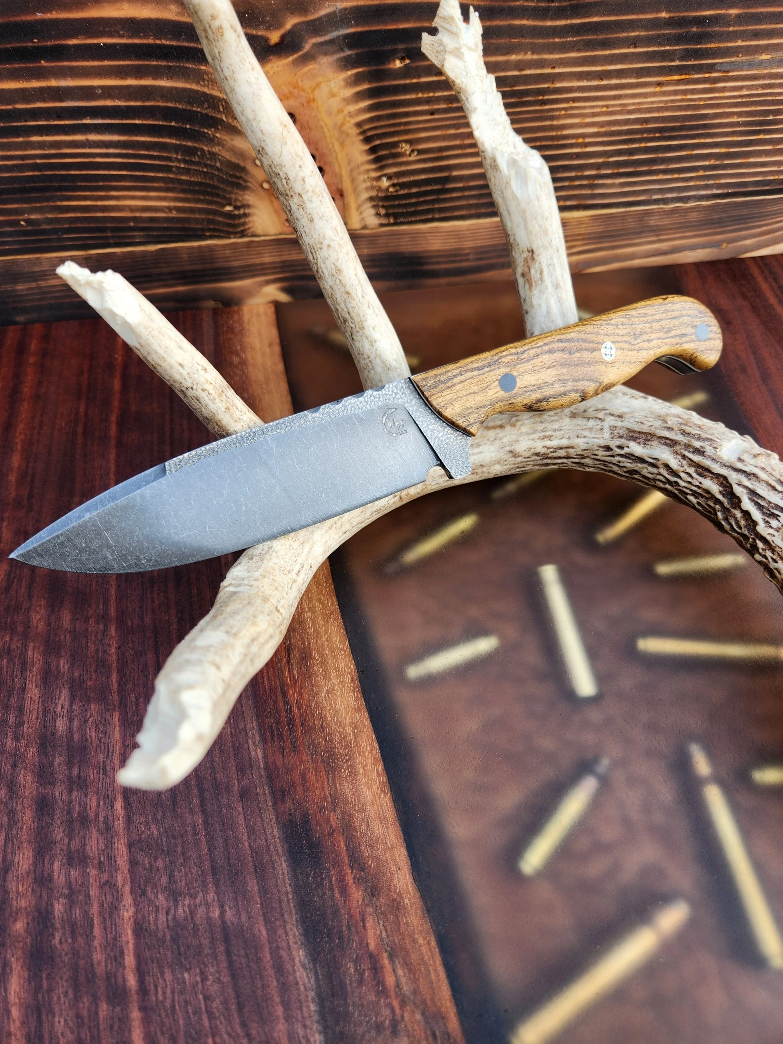 A knife with a wooden handle and a textured metal blade resting on a piece of driftwood, with a wooden clock in the background.