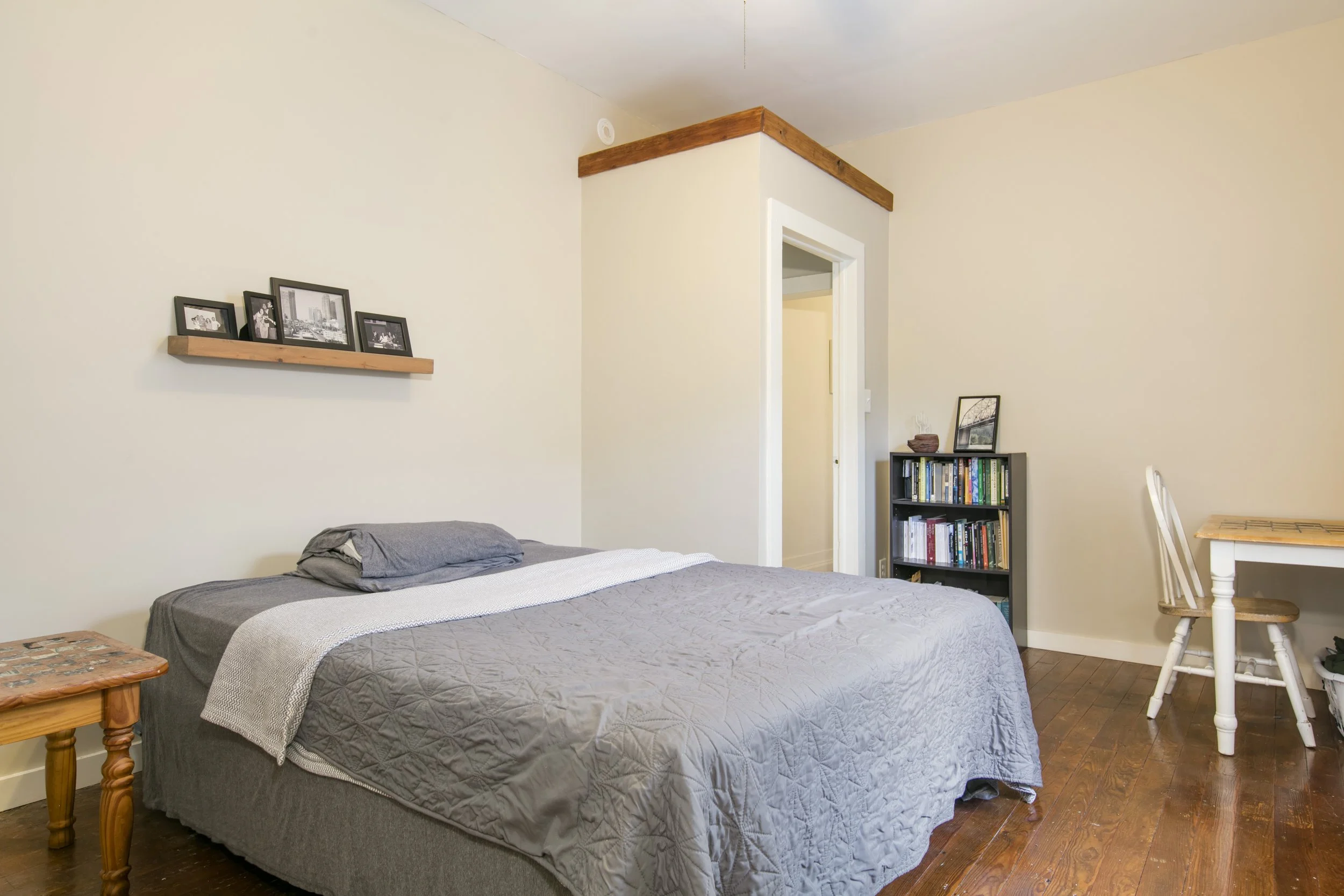 A bedroom with a bed covered in gray quilt and pillows, a nightstand on the left, a wall shelf with framed photographs, a bookshelf filled with books, a framed picture on top, and a white wooden chair.