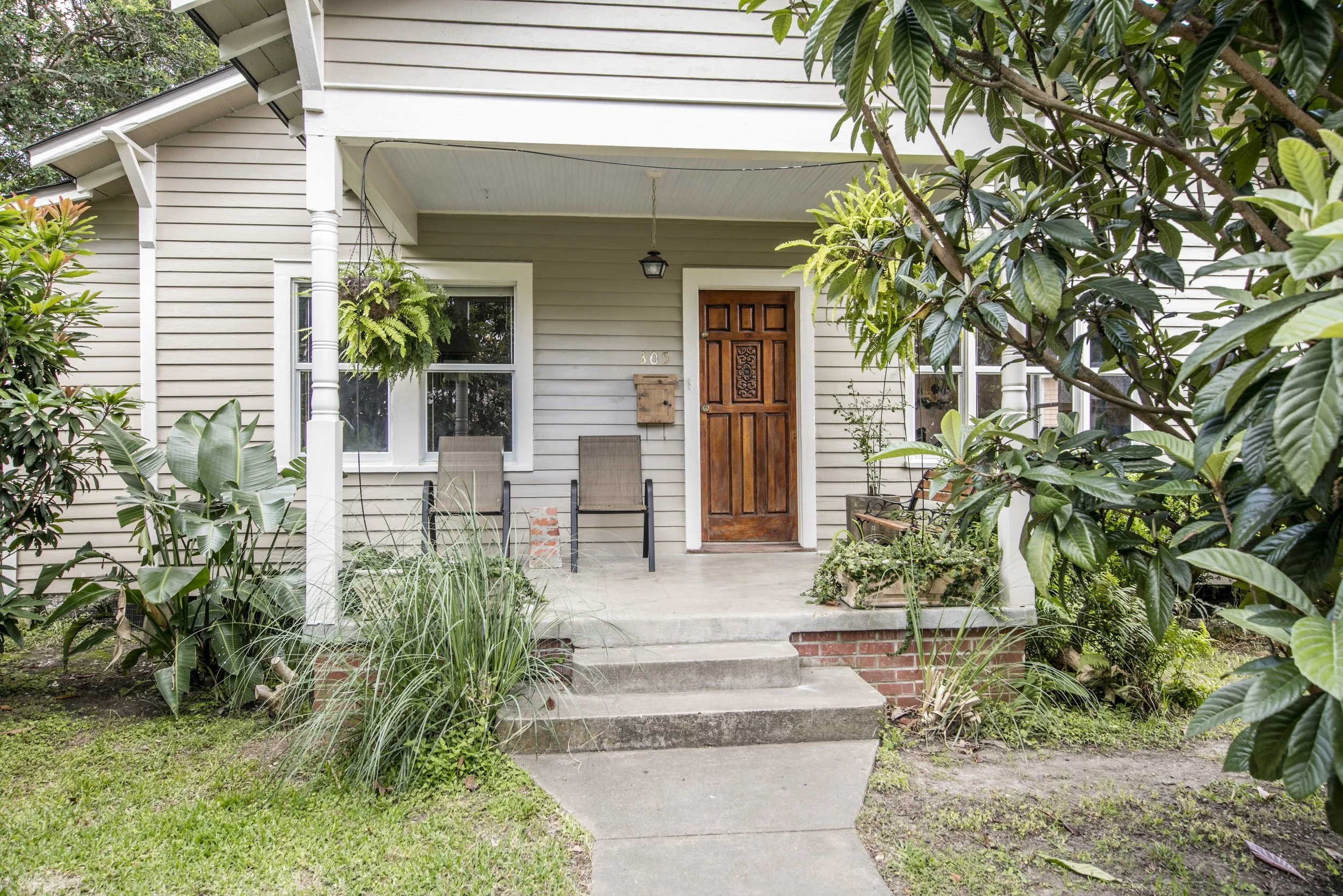 Front porch of a house with a wooden front door, two chairs, and lush greenery and plants surrounding the steps leading up to the porch.