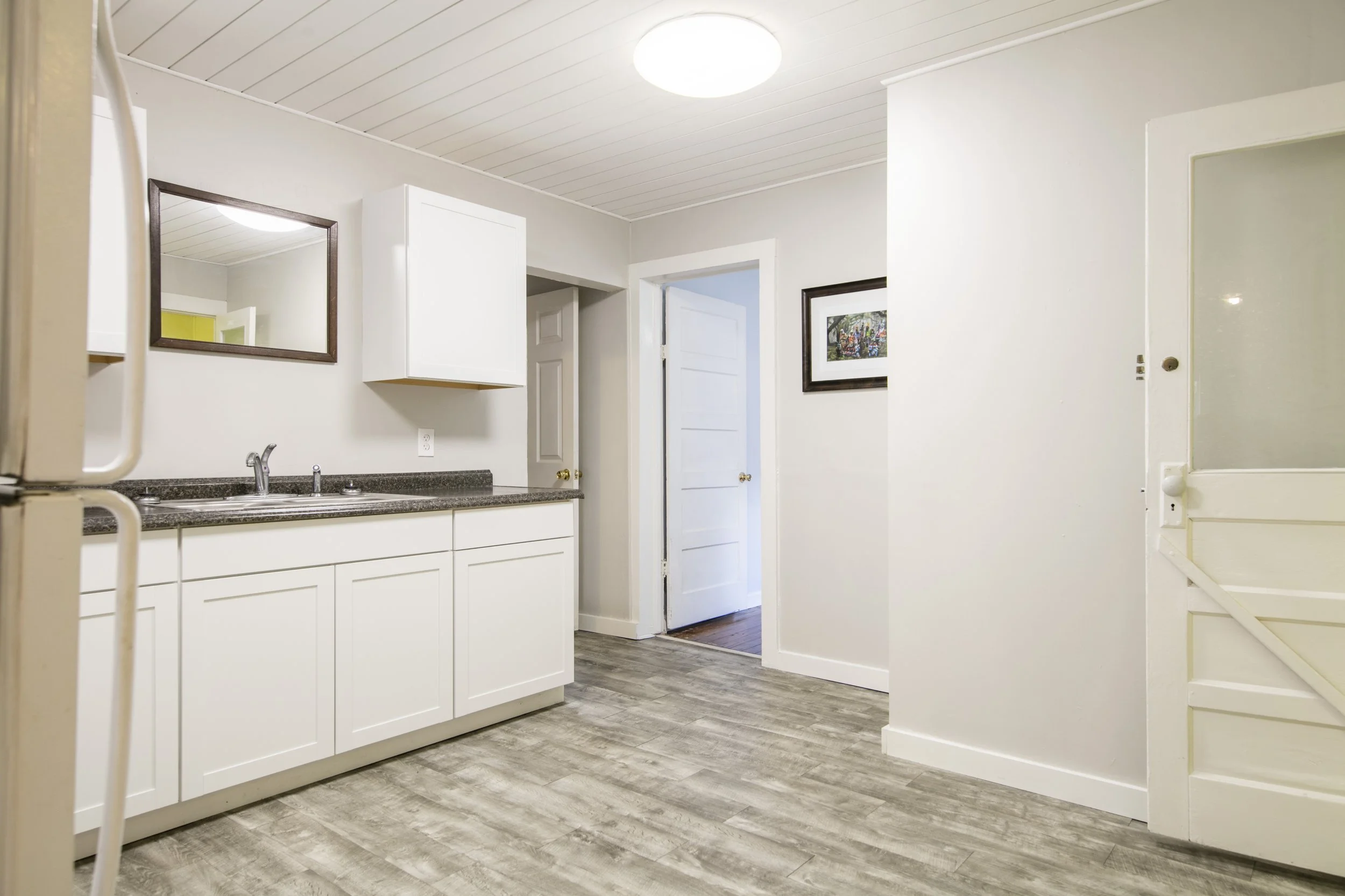 A kitchen with white cabinets, a granite countertop, a stainless steel sink, and a mirror on the wall. There is a door open to another room and a framed picture on the wall. The floor has wood-like gray tiles, and the ceiling has white beadboard paneling with a round ceiling light.