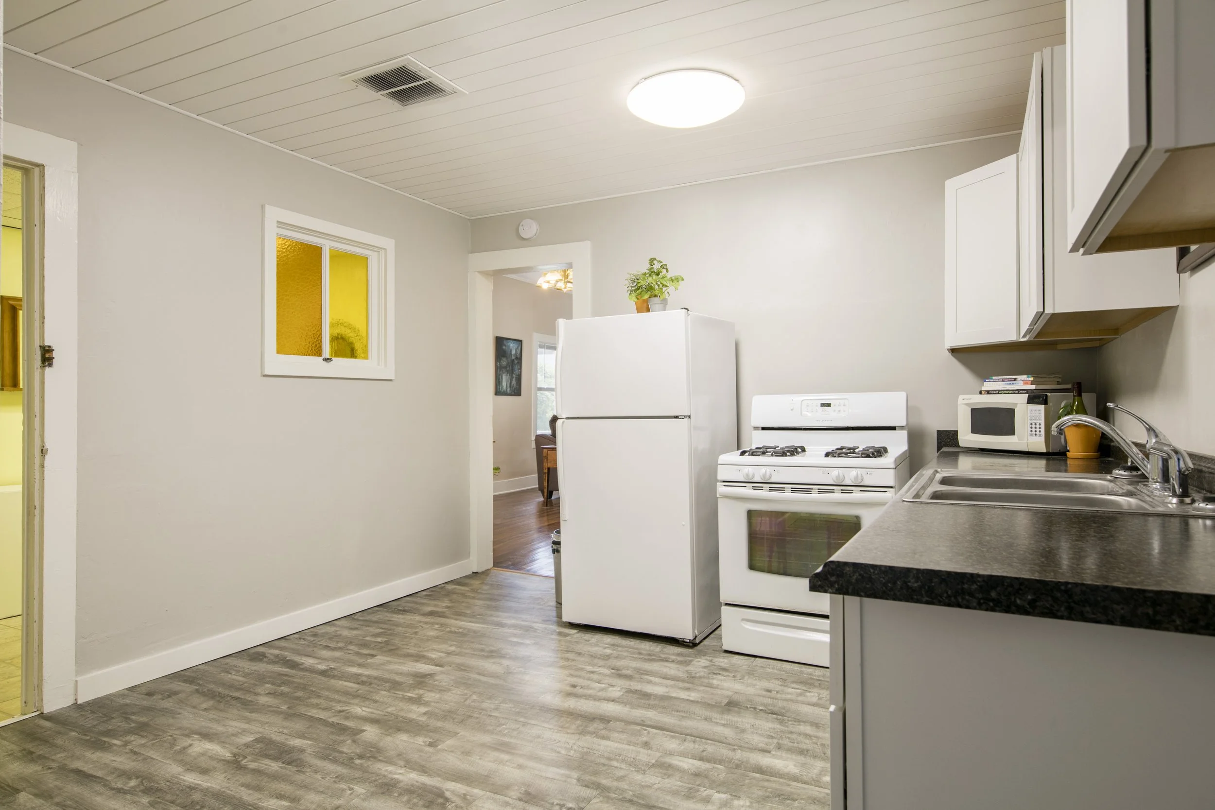 A kitchen with white walls, white cabinets, a white refrigerator, stove, microwave, and stainless steel sink, with a window and wooden floors.
