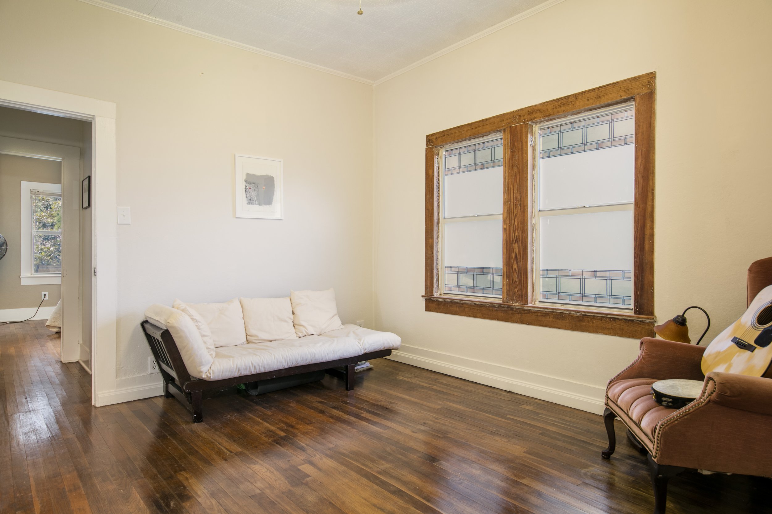 A cozy living room with hardwood floors, beige walls, a sofa with white cushions, a decorative framed artwork, and a window with wooden trim.