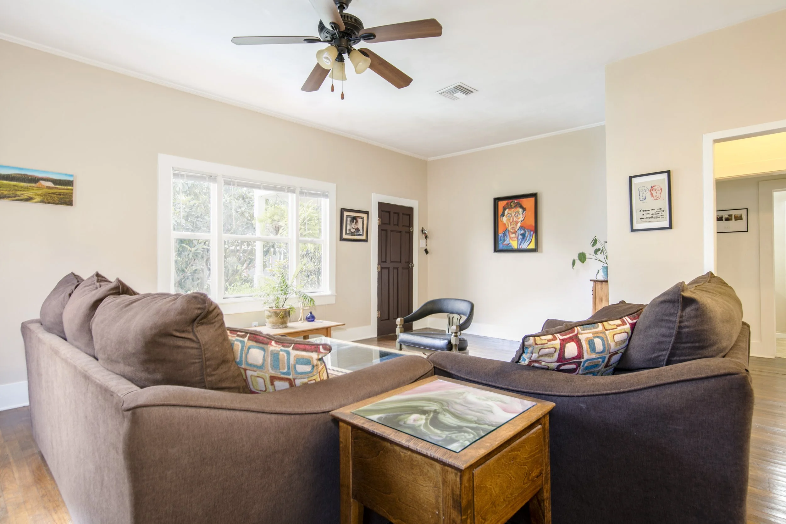 Living room with a beige sofa, colorful patterned pillows, glass-topped coffee table, black armchair, large window, and artwork on the walls.