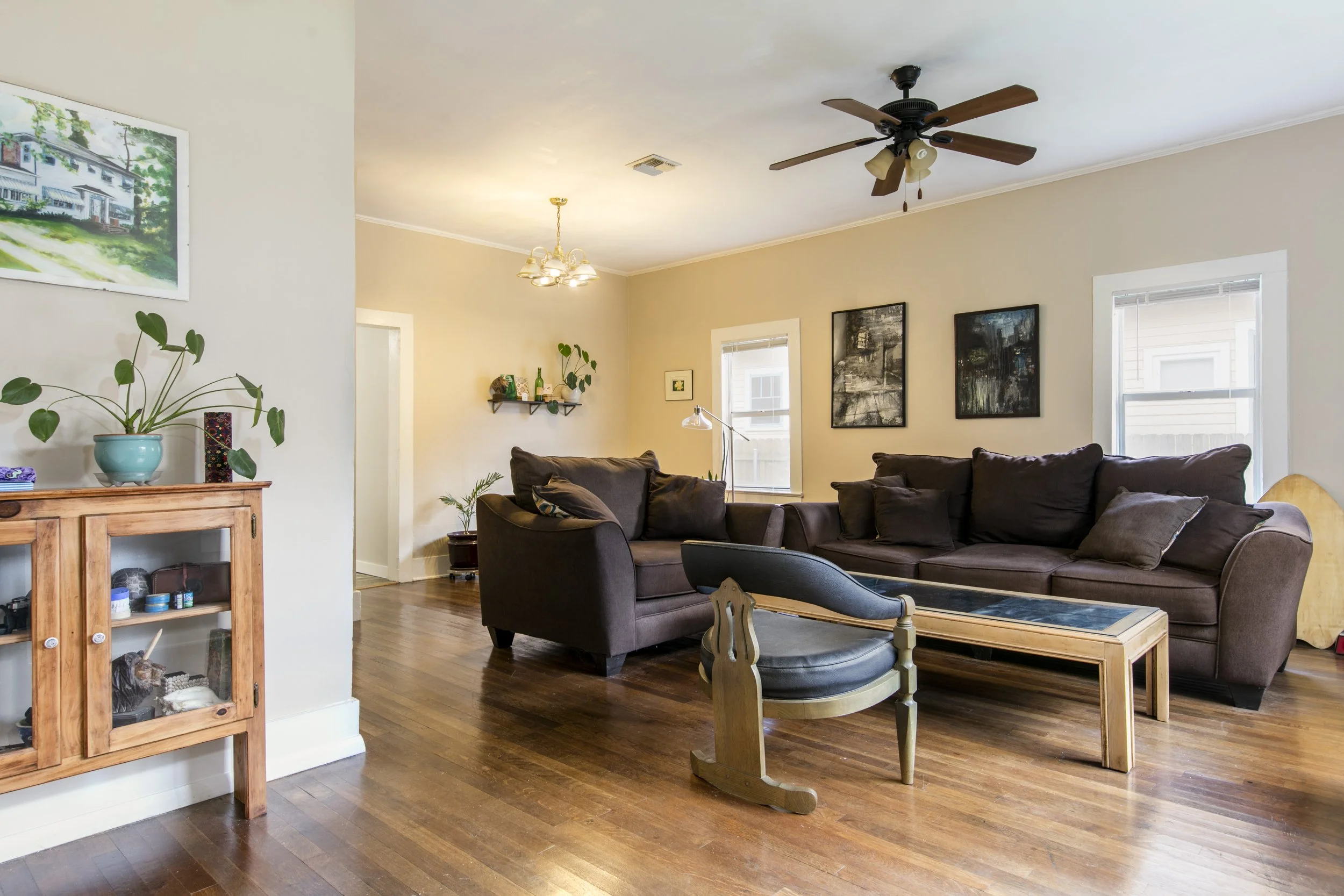 Living room with beige walls, hardwood floors, a dark brown sectional sofa, a wooden coffee table, artwork on the walls, and a ceiling fan with lights.