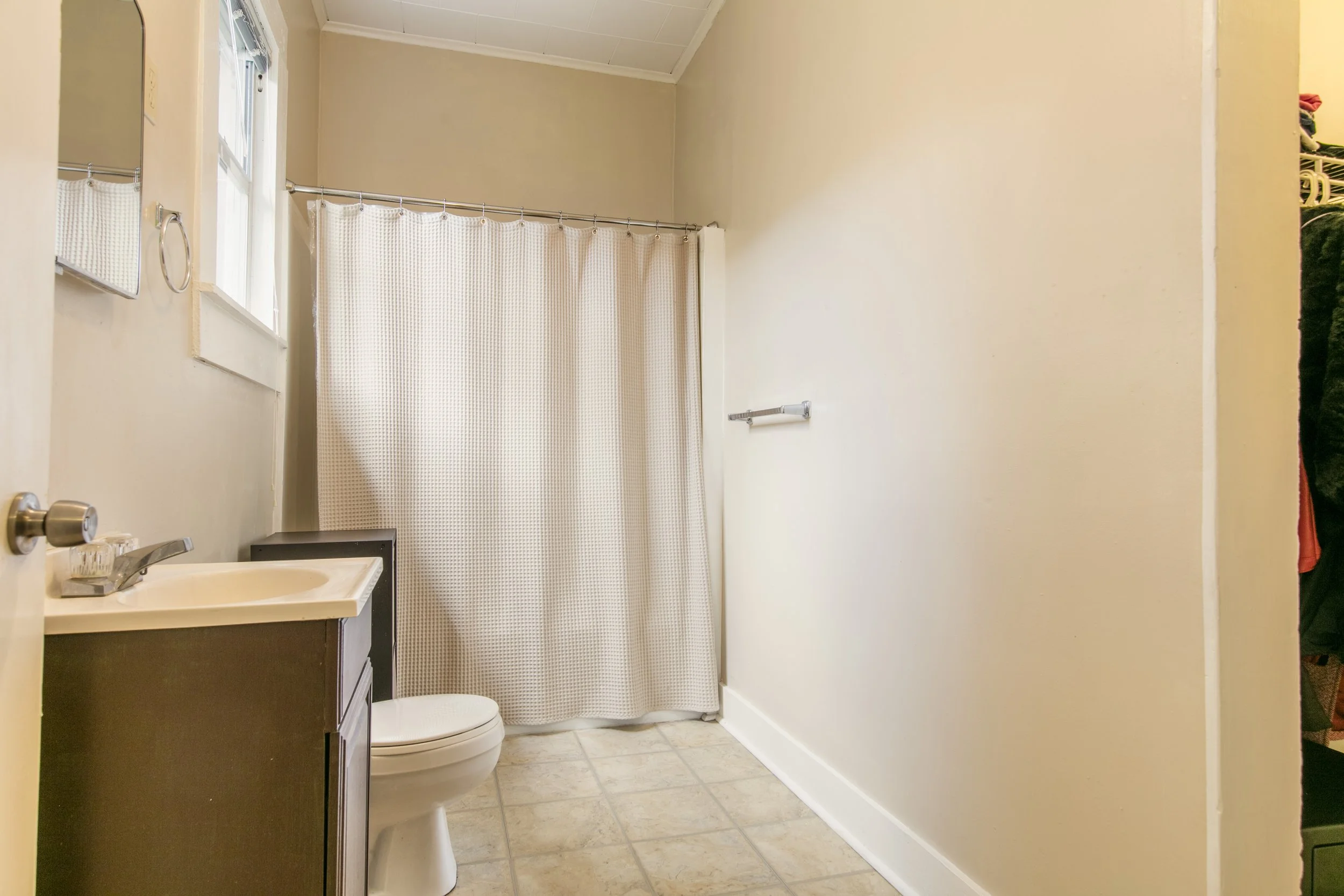 This is a small bathroom with a sink, mirror, toilet, and a shower with a beige curtain. The room has beige walls, light-colored tiled floor, and small windows above the shower.