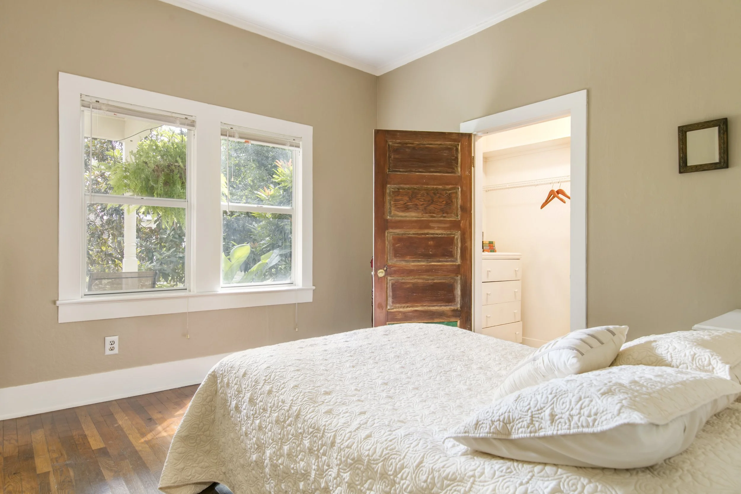 A bedroom with two twin beds covered with white quilted bedspreads, beige walls, hardwood floor, and a window with white trim showing greenery outside. An open wooden door reveals a closet with white drawers and pink hangers.