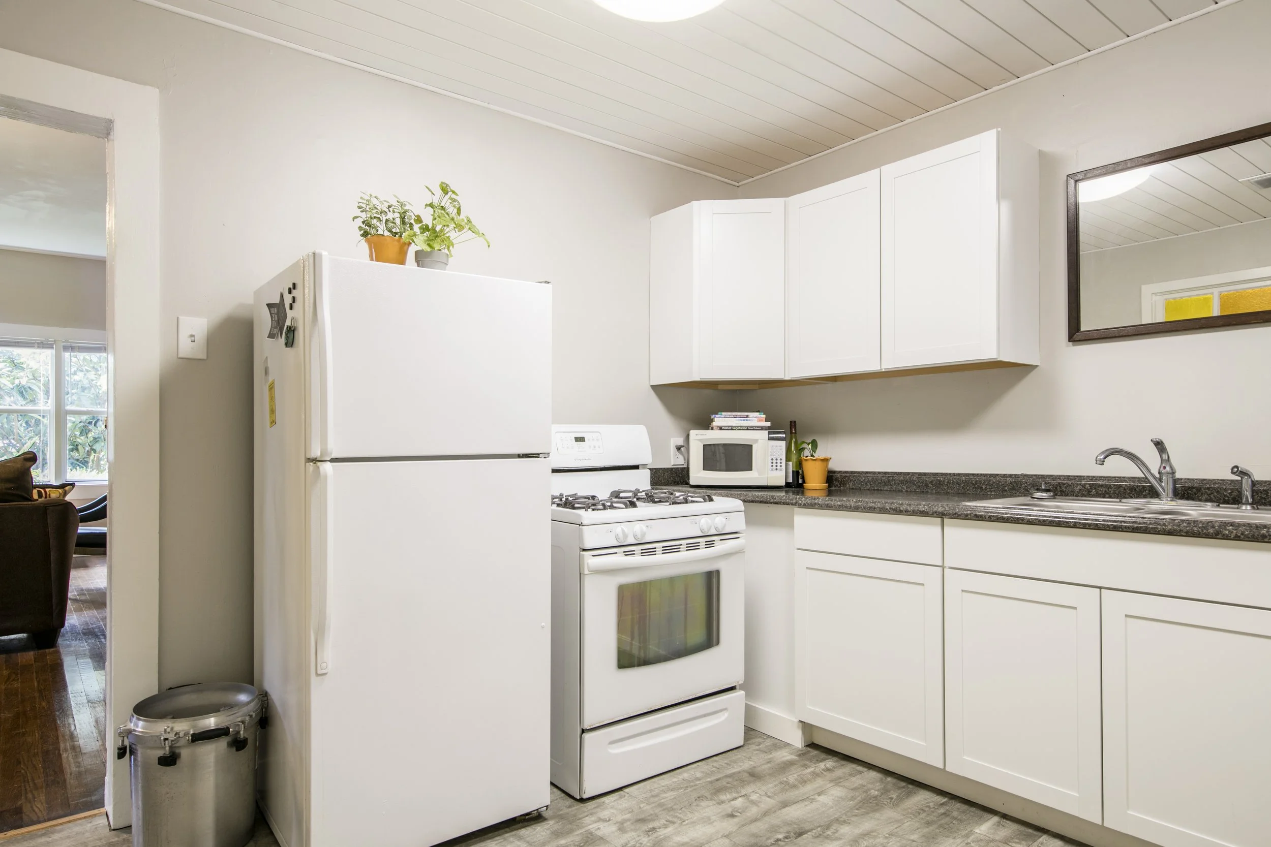 A kitchen with white cabinets, a white stove, a microwave, and a white refrigerator with potted plants on top. There is a mirror on the wall above the countertop, and the kitchen is adjacent to a living room area with a window showing trees outside.