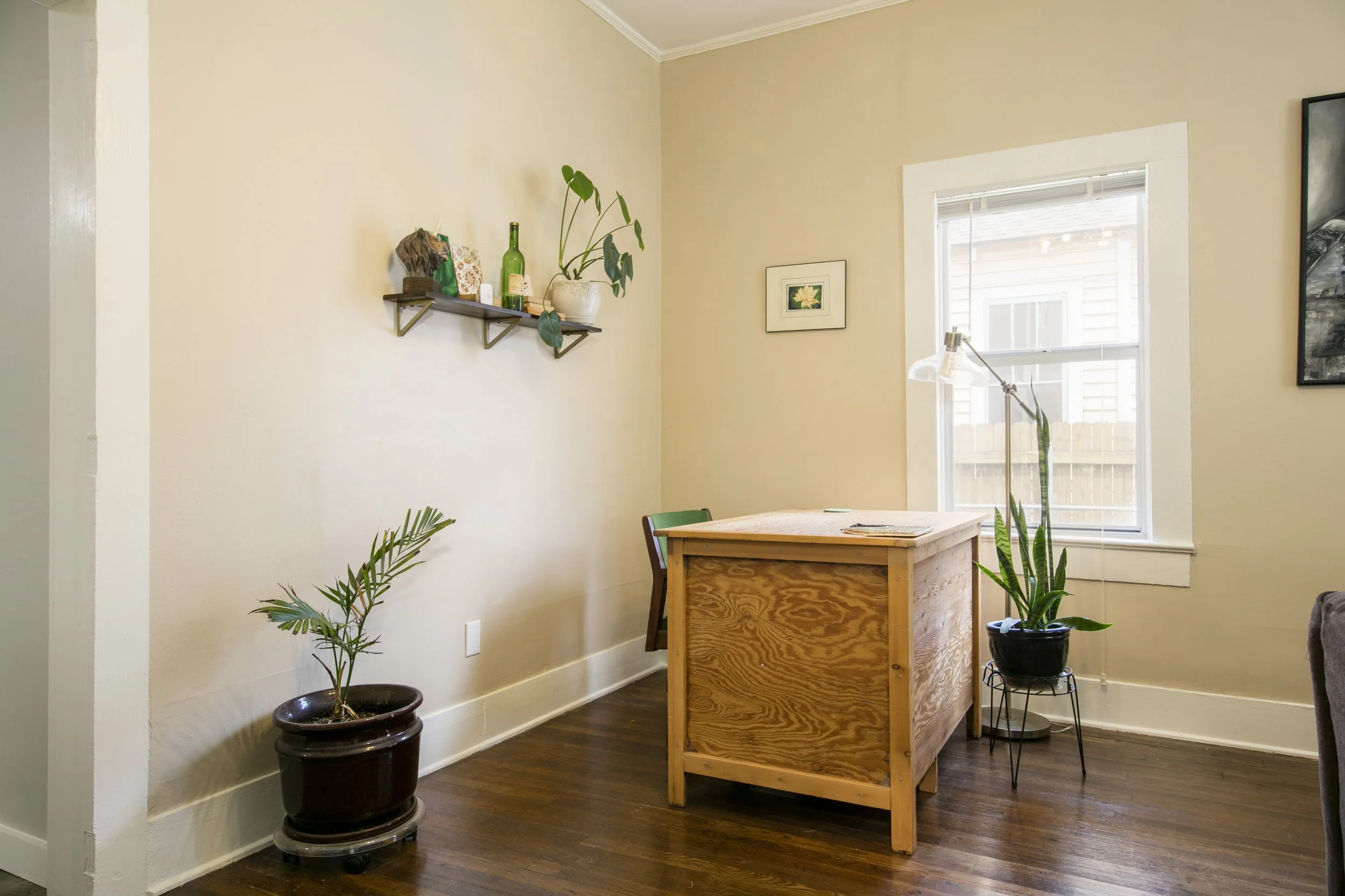 A cozy dining area with a wooden table, a green chair, potted plants, a wall shelf with decorative items, and a window with blinds.