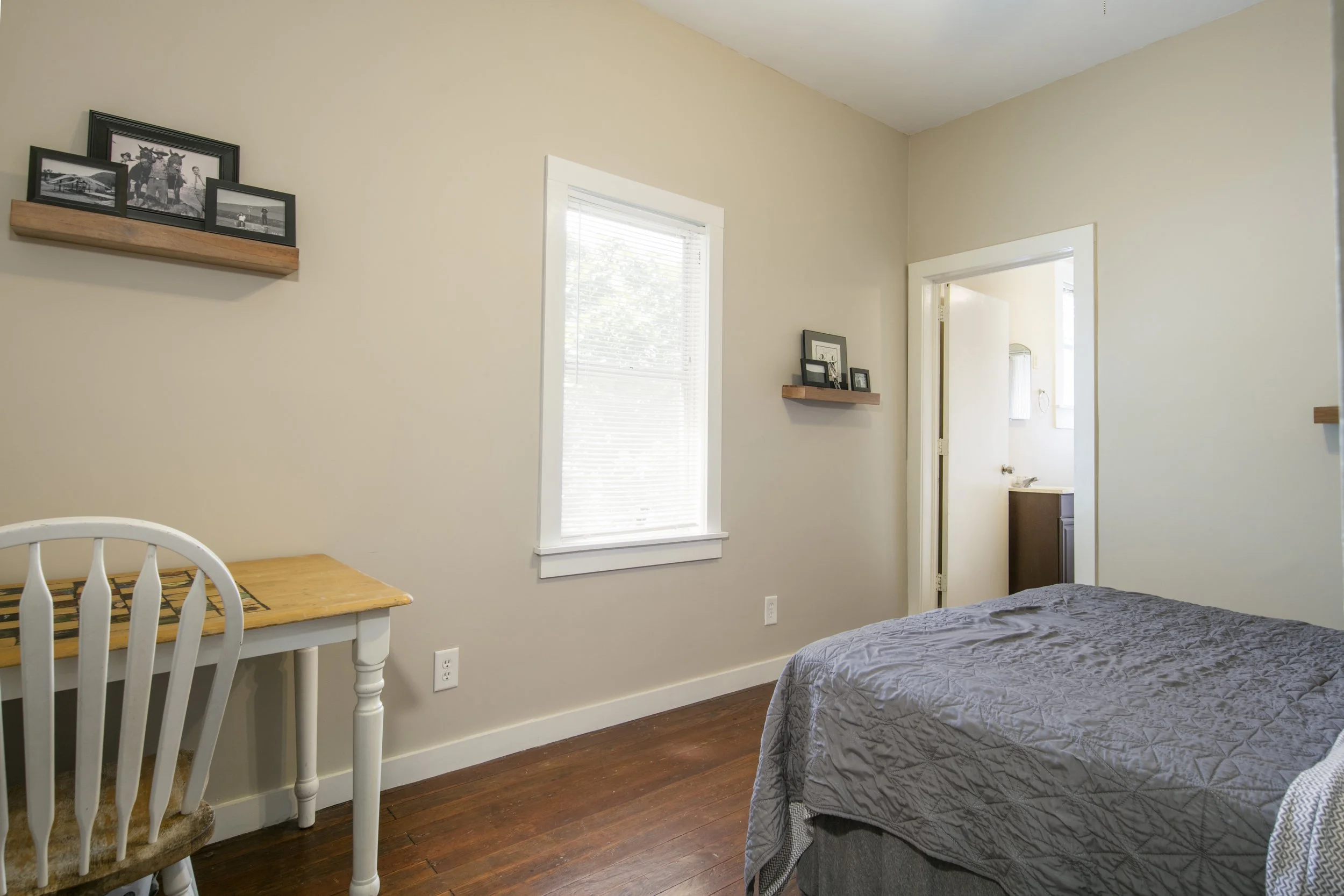 A bedroom with beige walls, a large window with blinds, a wooden shelf with framed photos, a bed with a gray quilt, a small desk with a chair, and hardwood flooring.