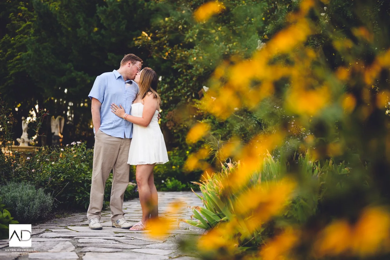 New Castle County engagement photographer captures couple kissing on stone path with yellow flowers and stone urn
