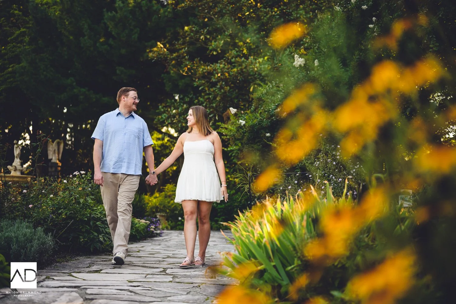 Couple walking hand-in-hand on stone path past black-eyed Susans and garden statue — New Castle County wedding photographer