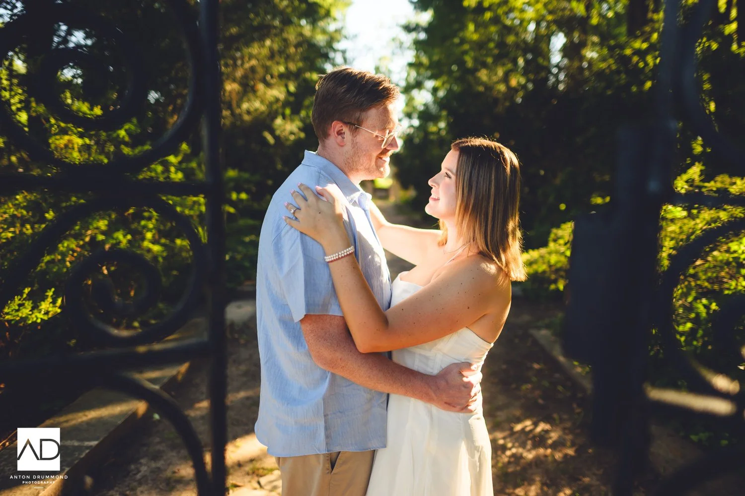Couple embracing by ornate iron garden gate, engagement ring visible — New Castle County engagement photographer