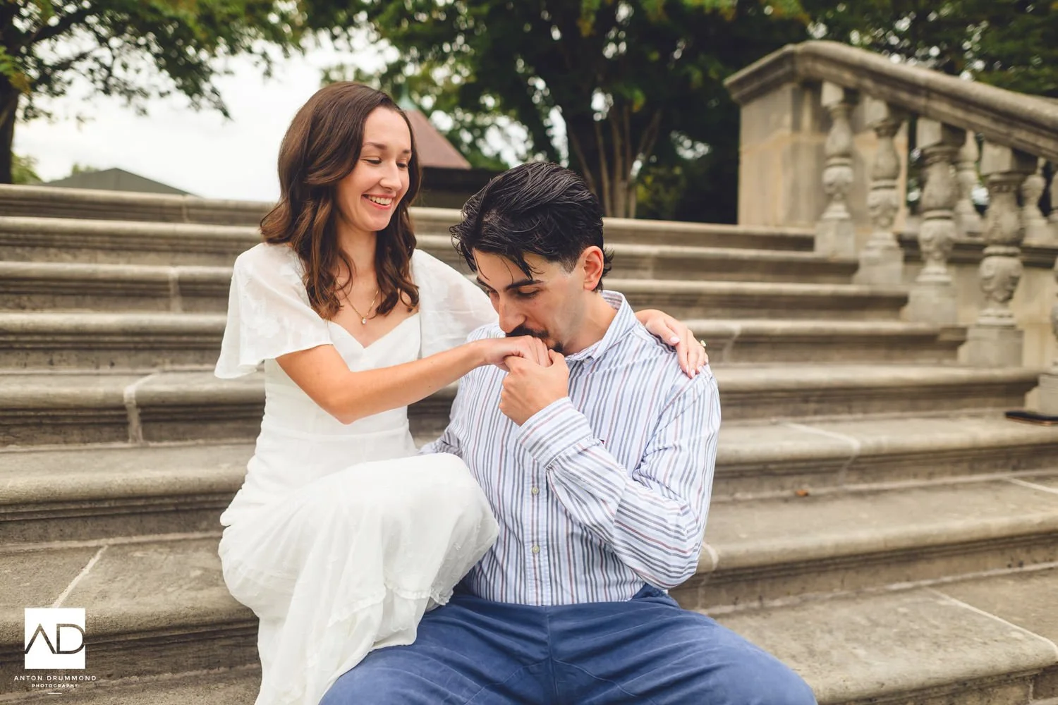 Joe kissing Emily's hand on the stone steps as she laughs and leans into him — Curtis Arboretum engagement session