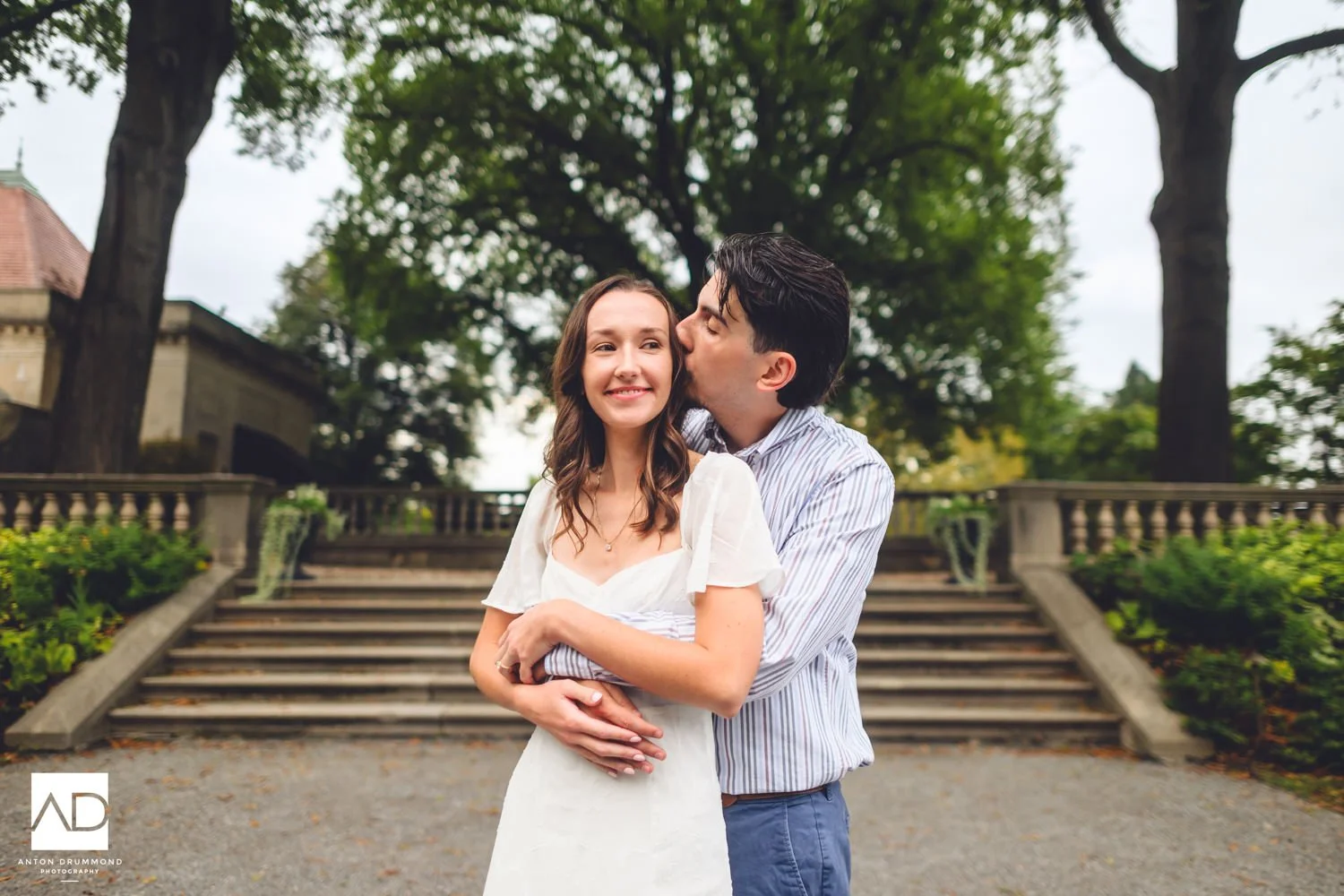 Anton Drummond captures Joe kissing Emily's cheek as she smiles at the camera, stone balustrade and trees behind