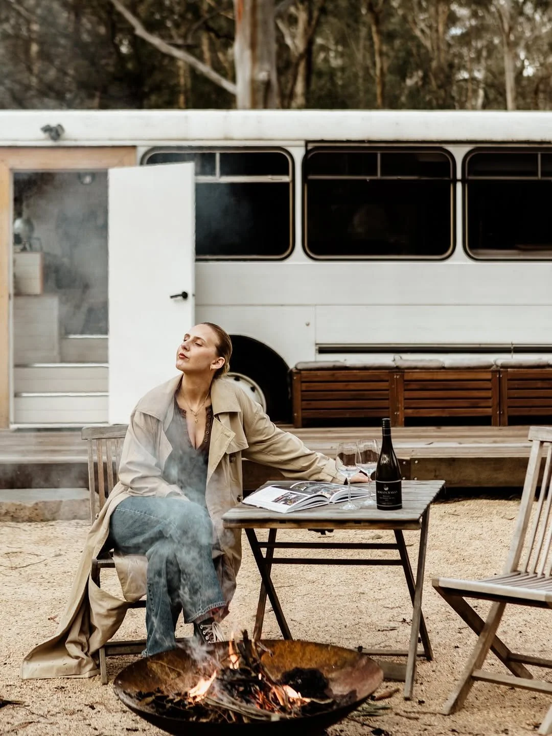 Woman relaxing by a fire pit with a glass of red wine outside the Moss and Wheels converted bus, Cottles Bridge Victoria