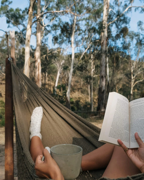 Guest relaxing in hammock with book and coffee cup surrounded by eucalyptus trees at Moss & Wheels bus accommodation Victoria