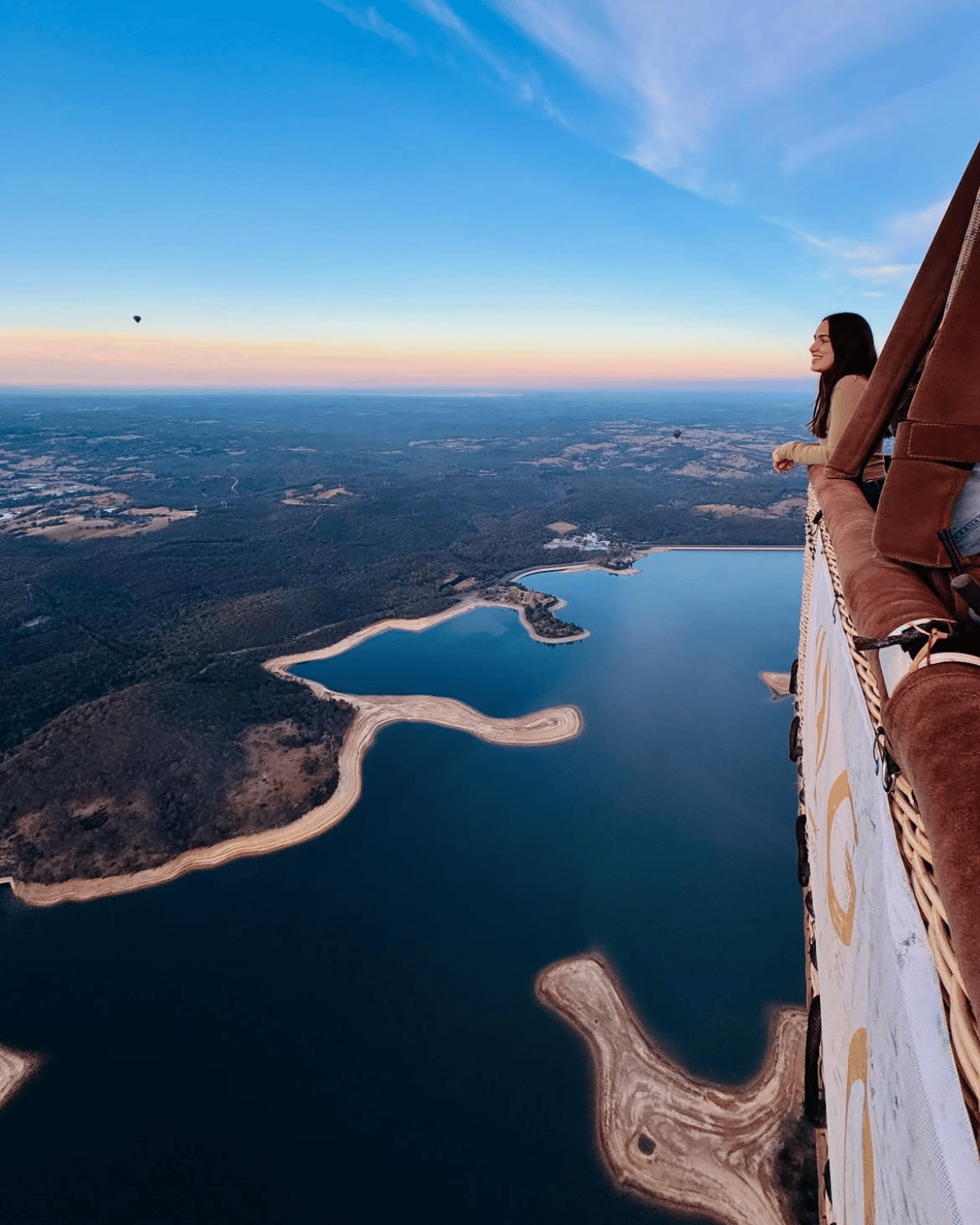 Woman looking out from a hot air balloon basket over a vast Sugarloaf Reservoir and treetops at sunrise in the Yarra Valley