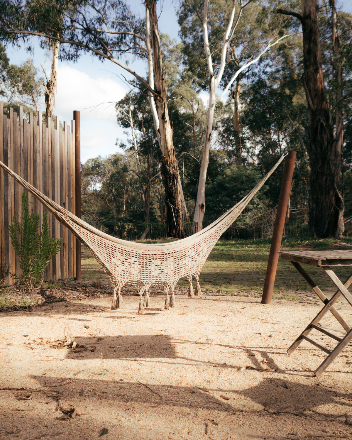 Macrame hammock with tassel fringe strung between wooden posts beside a timber fence at Moss & Wheels Cottles Bridge