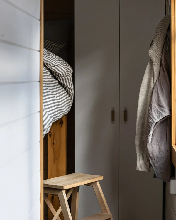 Cosy interior of Moss & Wheels converted bus accommodation showing timber step stool, taupe wardrobe, linen layers and elevated queen bed — unique stay in Victoria's countryside