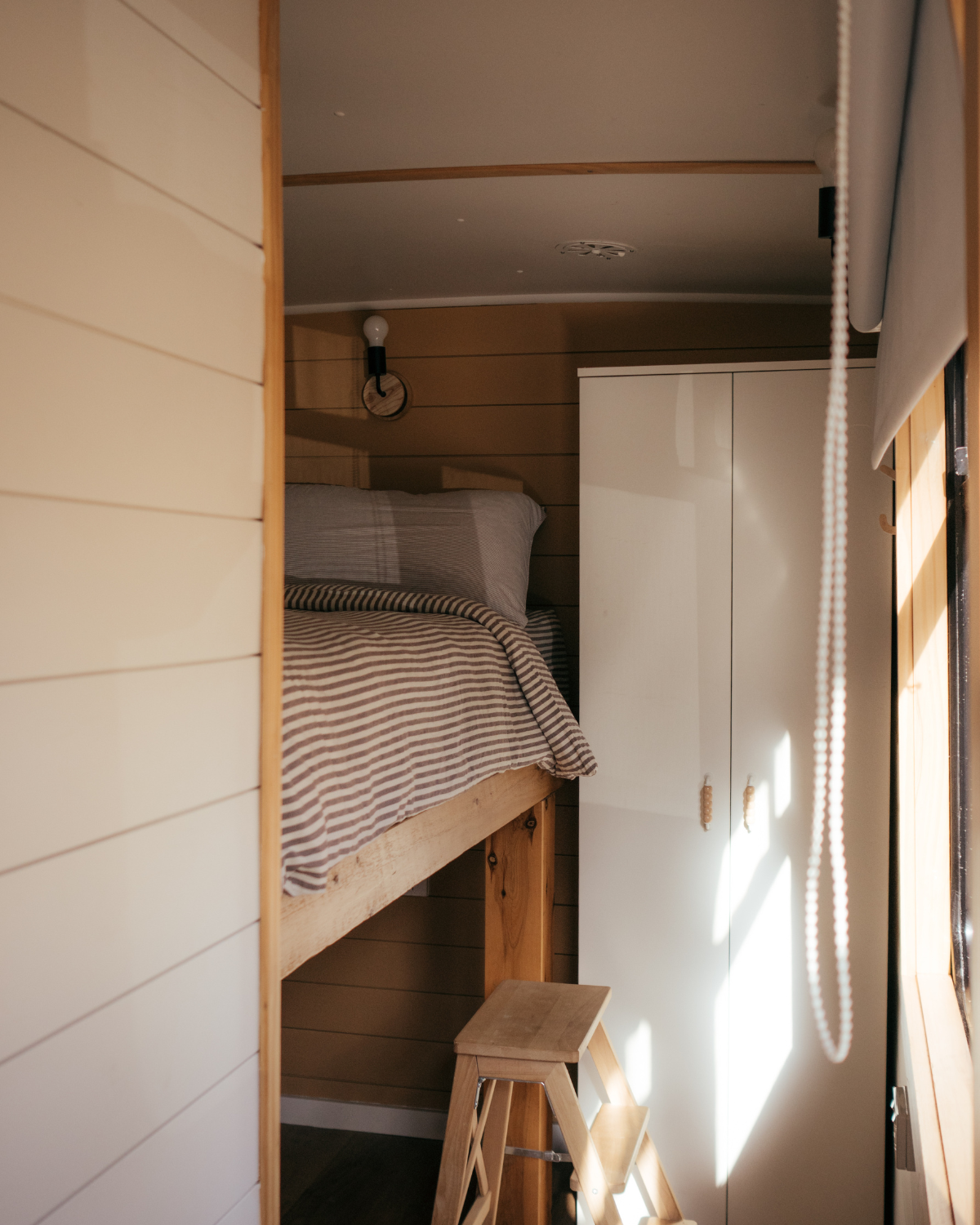 Cosy elevated bed with striped linen and wooden step stool inside the Moss & Wheels converted bus bedroom