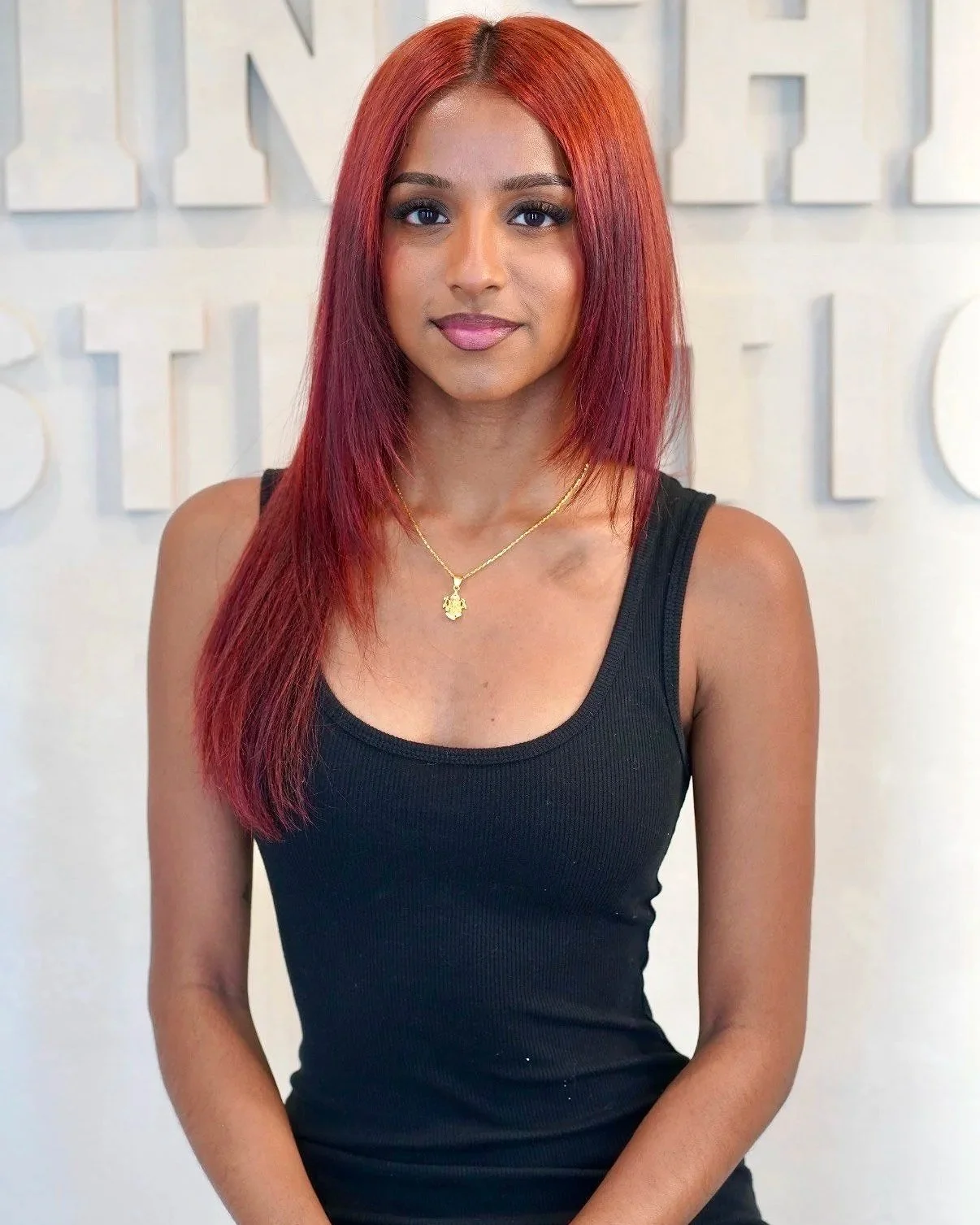 A woman with straight red hair, wearing a black tank top and a gold necklace, standing in front of a white background with partially visible large letters.