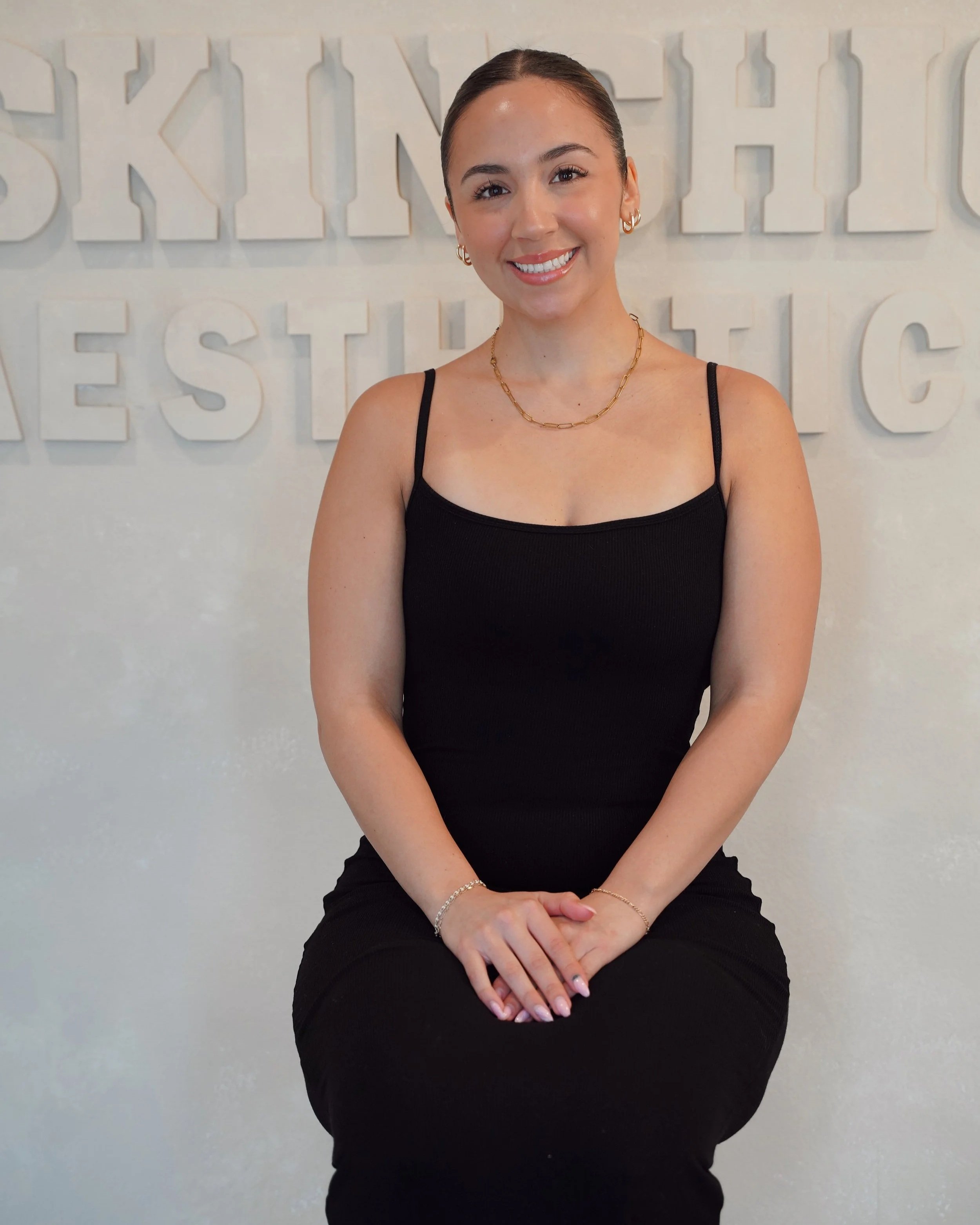 A young woman sitting against a white wall with large text, wearing a black spaghetti strap top, gold jewelry, and smiling.