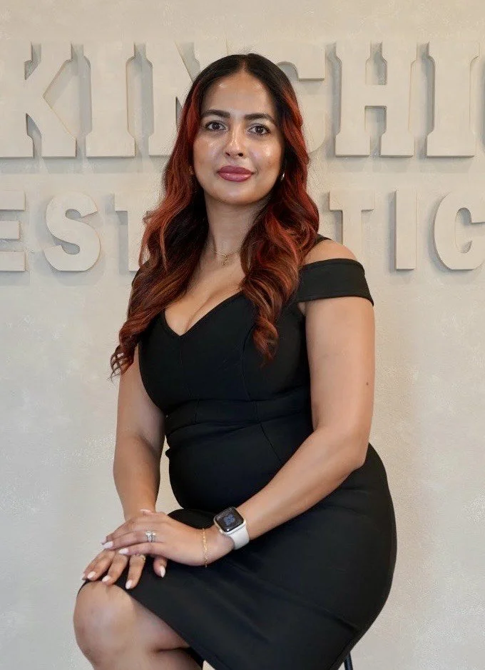 A pregnant woman with long, wavy red hair, wearing a fitted black dress with cold shoulder sleeves, sitting on a chair, and smiling at the camera, in front of a wall with large white decorative letters.