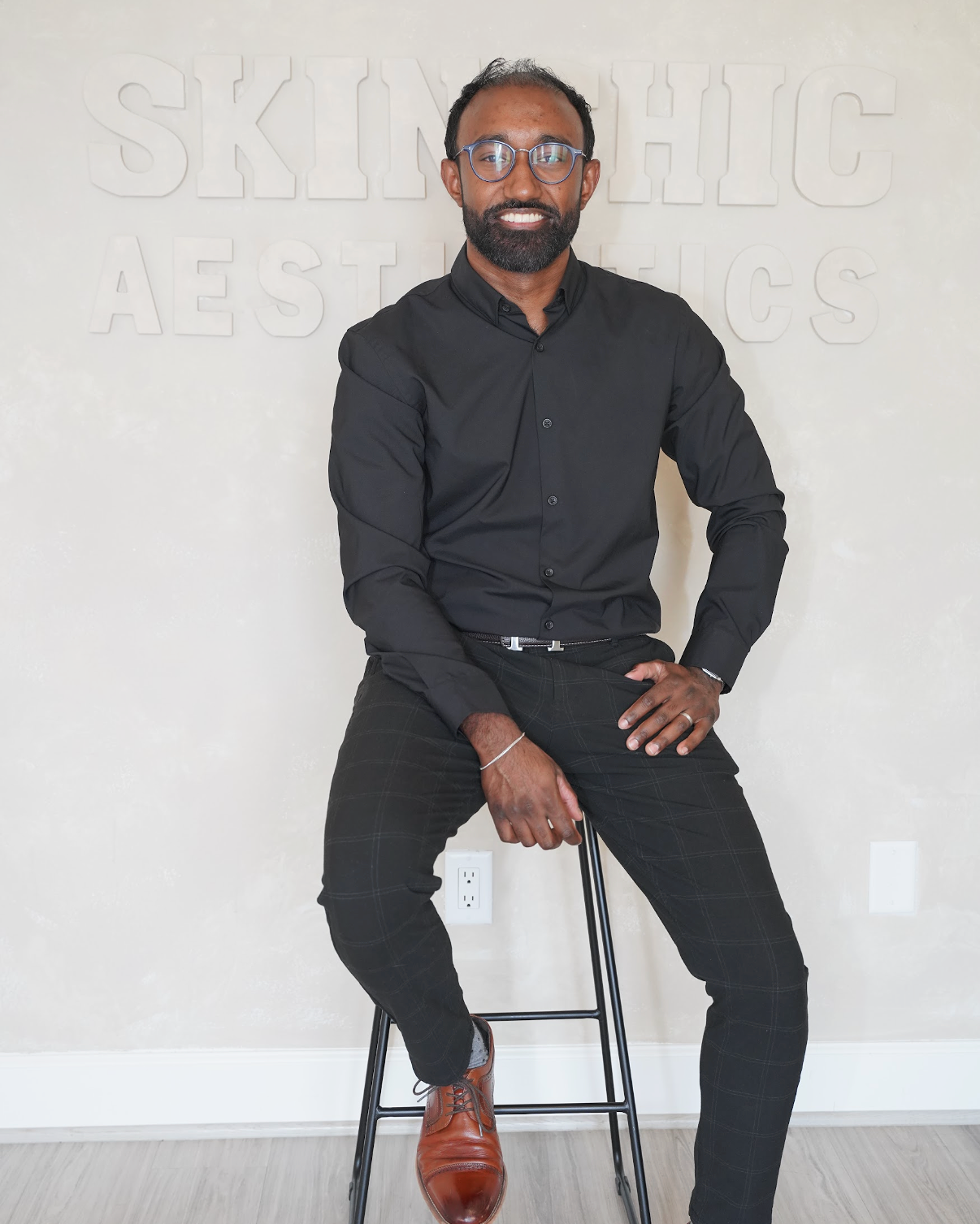 A man with glasses, a beard, and wearing a black shirt and plaid pants sitting on a stool in front of a wall with the partially visible text 'SKIN THIC AESTHETICS' in raised letters.