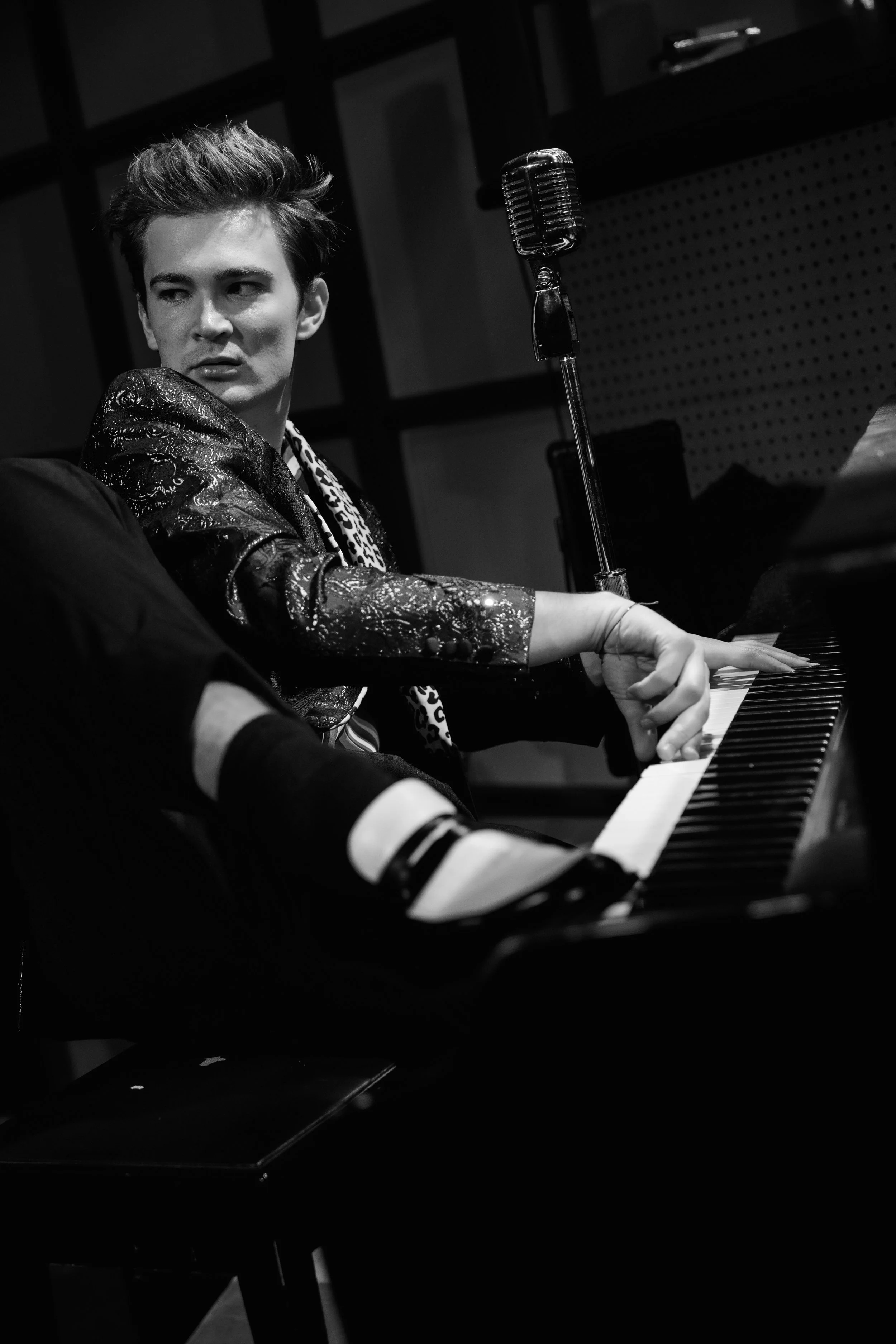 A black and white photo of a young man sitting at a grand piano, looking thoughtfully to the side, with a vintage microphone positioned in front of him.