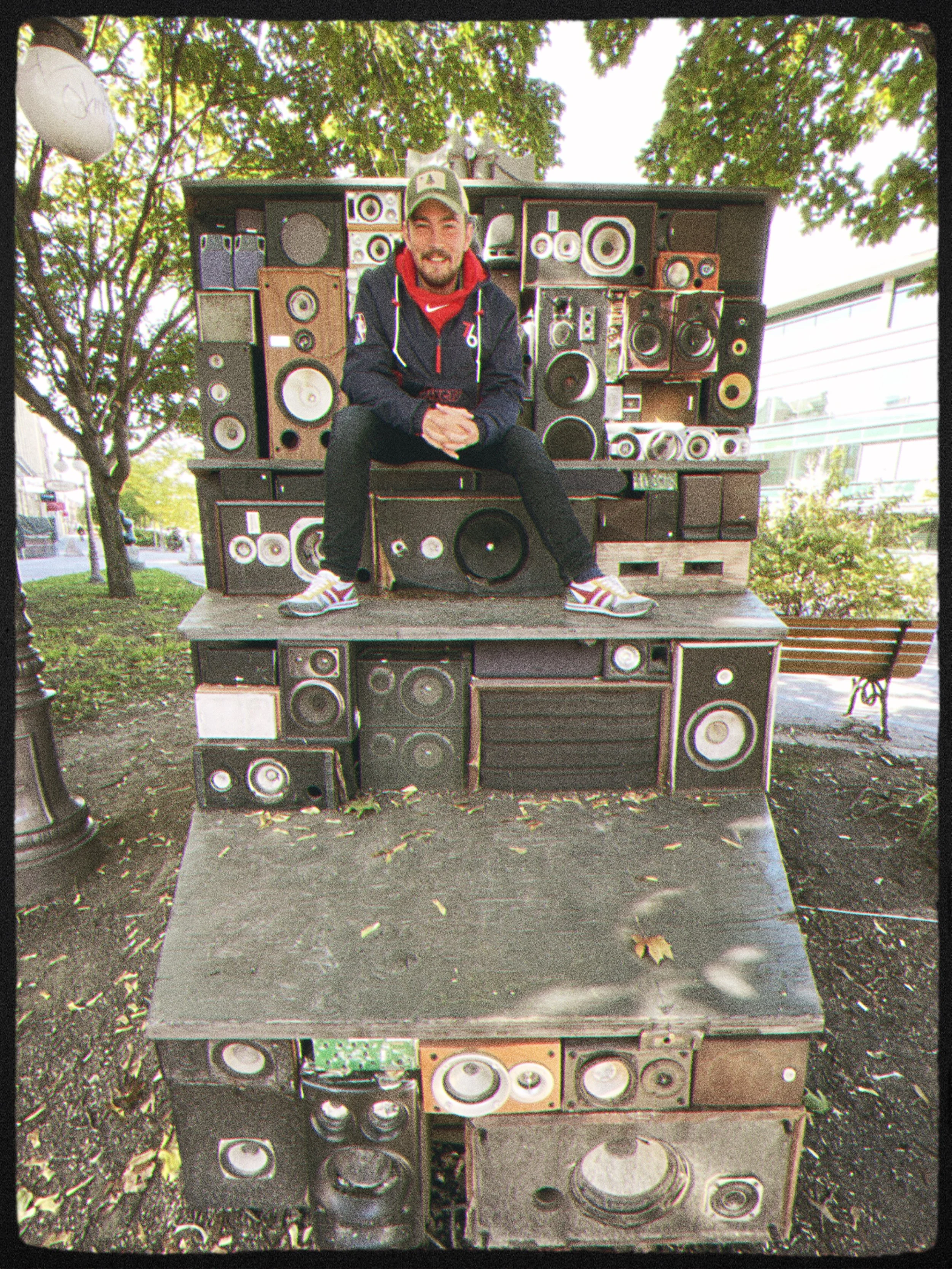 Giacomo sitting on a structure made of vintage speakers outdoors under a tree.