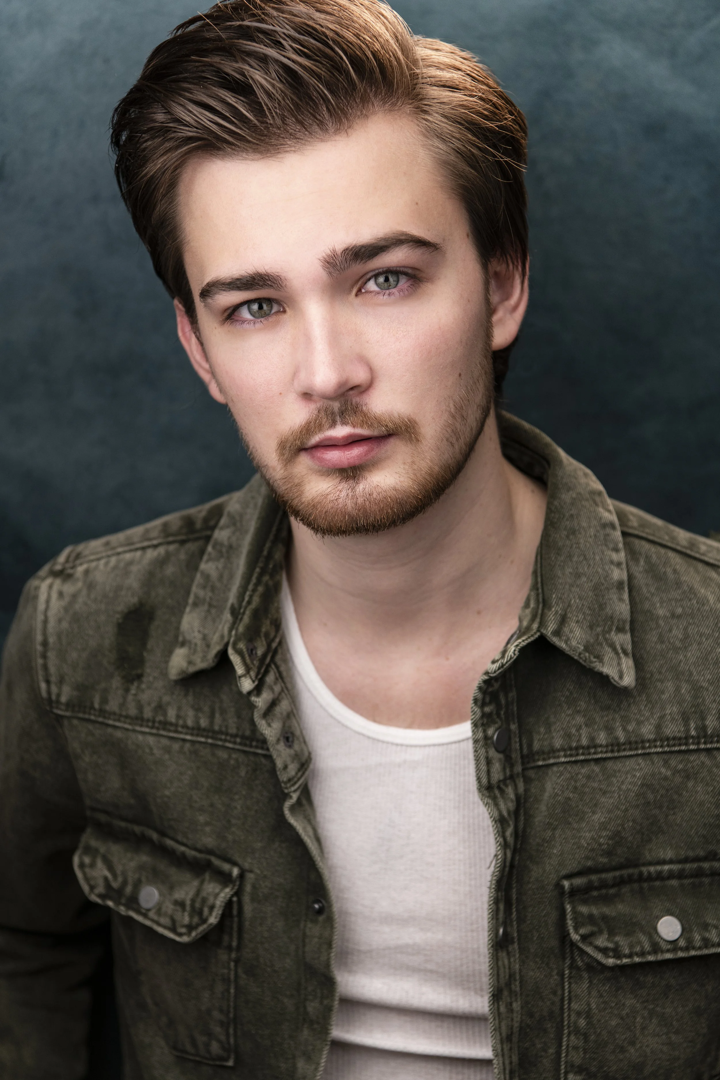 Giacomo with fair skin, light eyes, and brown hair is looking at the camera. He has a beard and is wearing a green military-style jacket over a white shirt, with a neutral expression. The background is dark green.