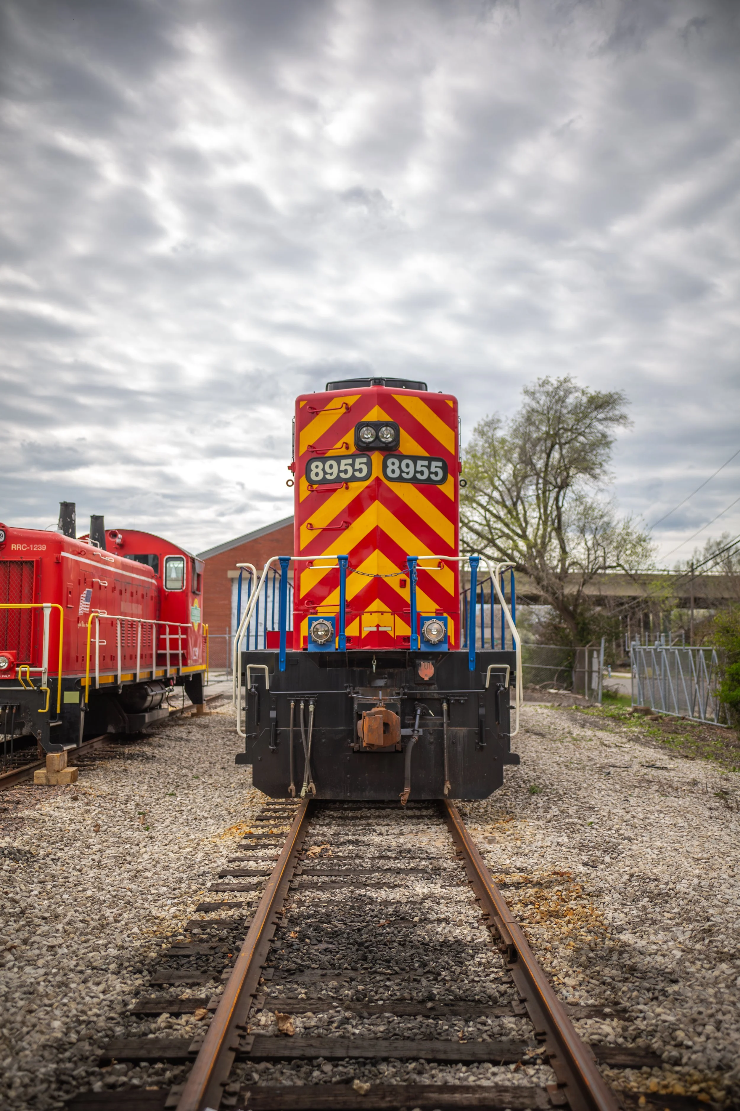 Front view of a red and yellow striped locomotive on train tracks with another red train car to the left and trees in the background.