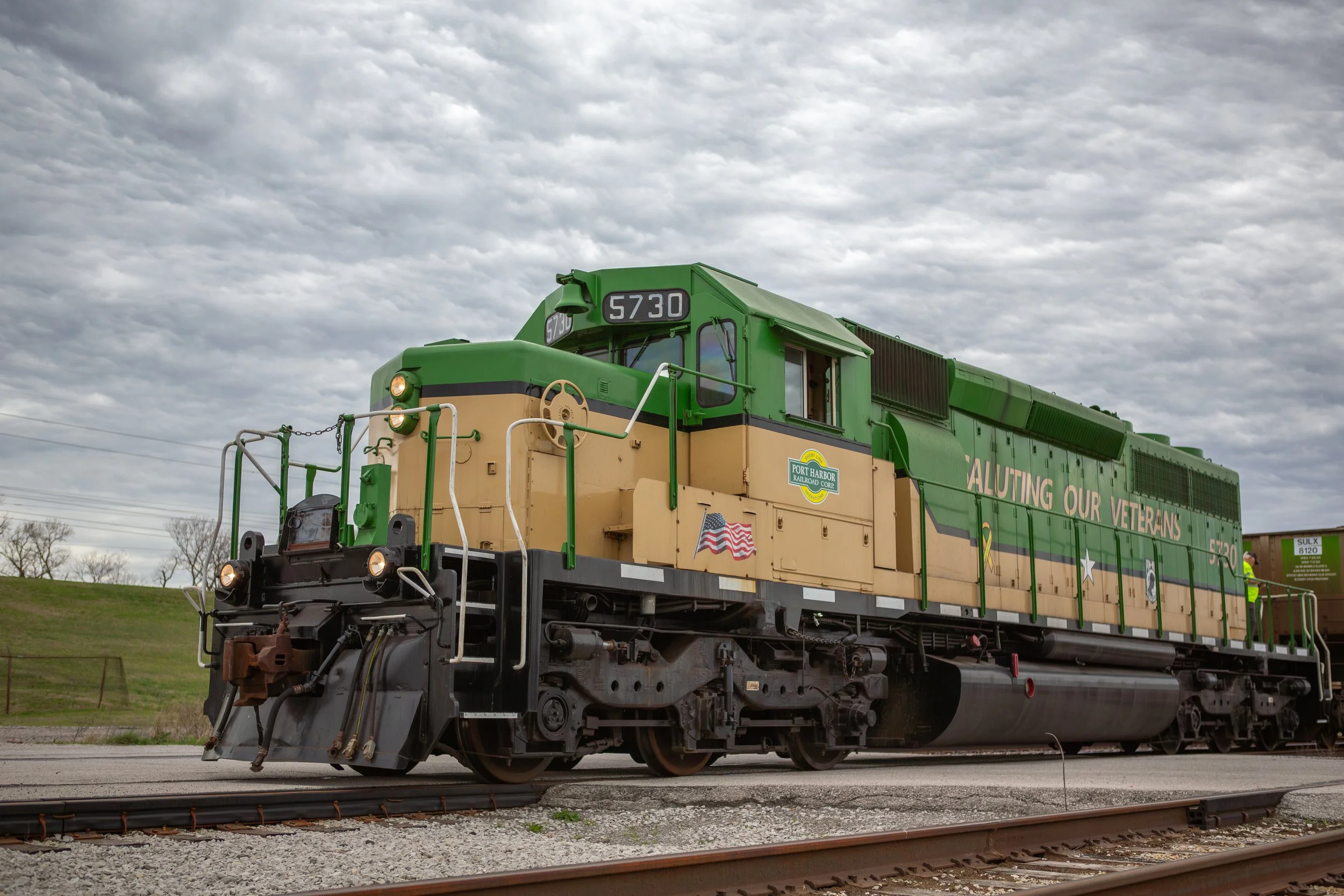 A green and tan locomotive train on tracks with a cloudy sky background, commemorating veterans with the message 'LUTING OUR VETERANS' and an American flag decal.