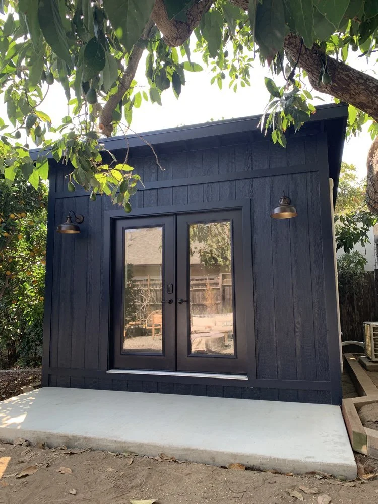 Small black shed with glass double doors, two wall-mounted exterior lights, surrounded by trees and greenery, on a concrete platform in a backyard.