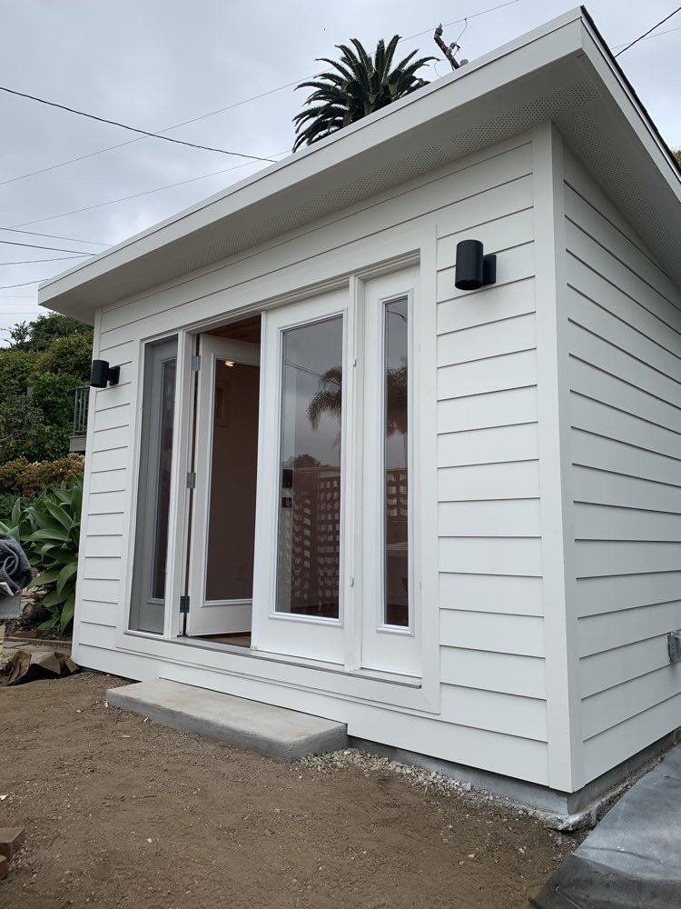 White modern small house or shed with sliding glass doors, two black exterior wall lights, surrounded by landscaping with plants and dirt, with a concrete pad in front.