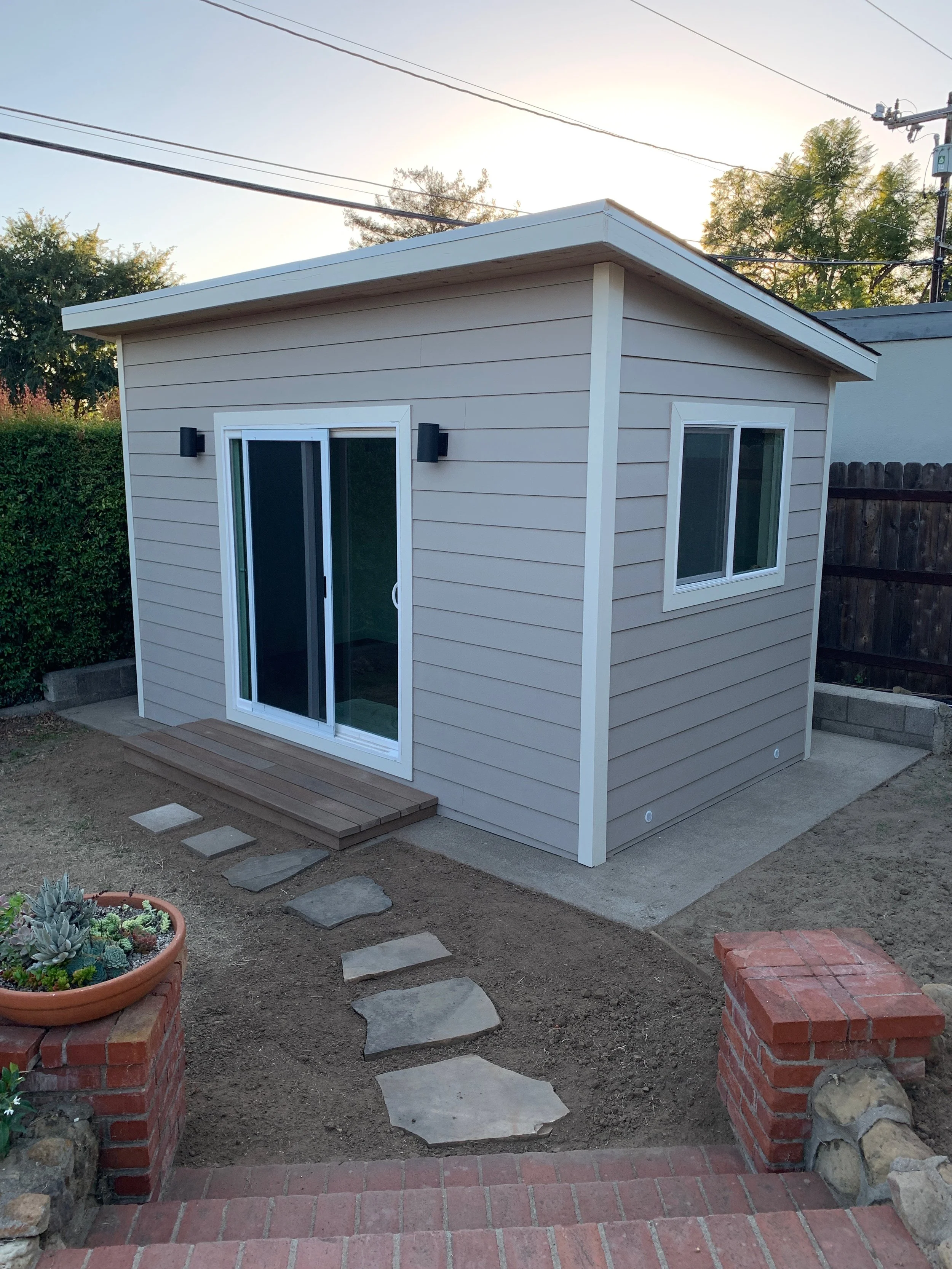 A small beige backyard studio shed with white trim and large sliding glass doors, two black exterior lights, situated on a backyard with a brick pathway, soil, and some plants, with trees and power lines in the background.