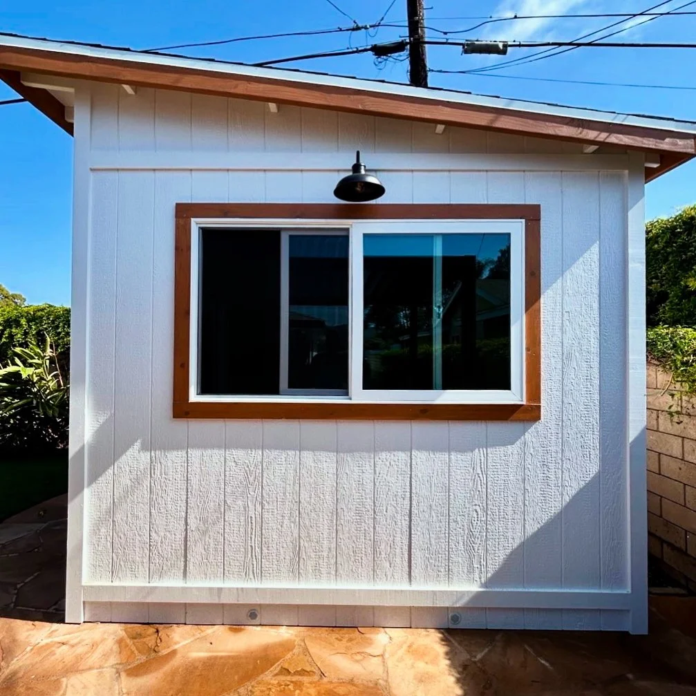 Small white backyard shed with a sliding window, wooden trim, black outdoor light fixture above the window, and a sloped roof.