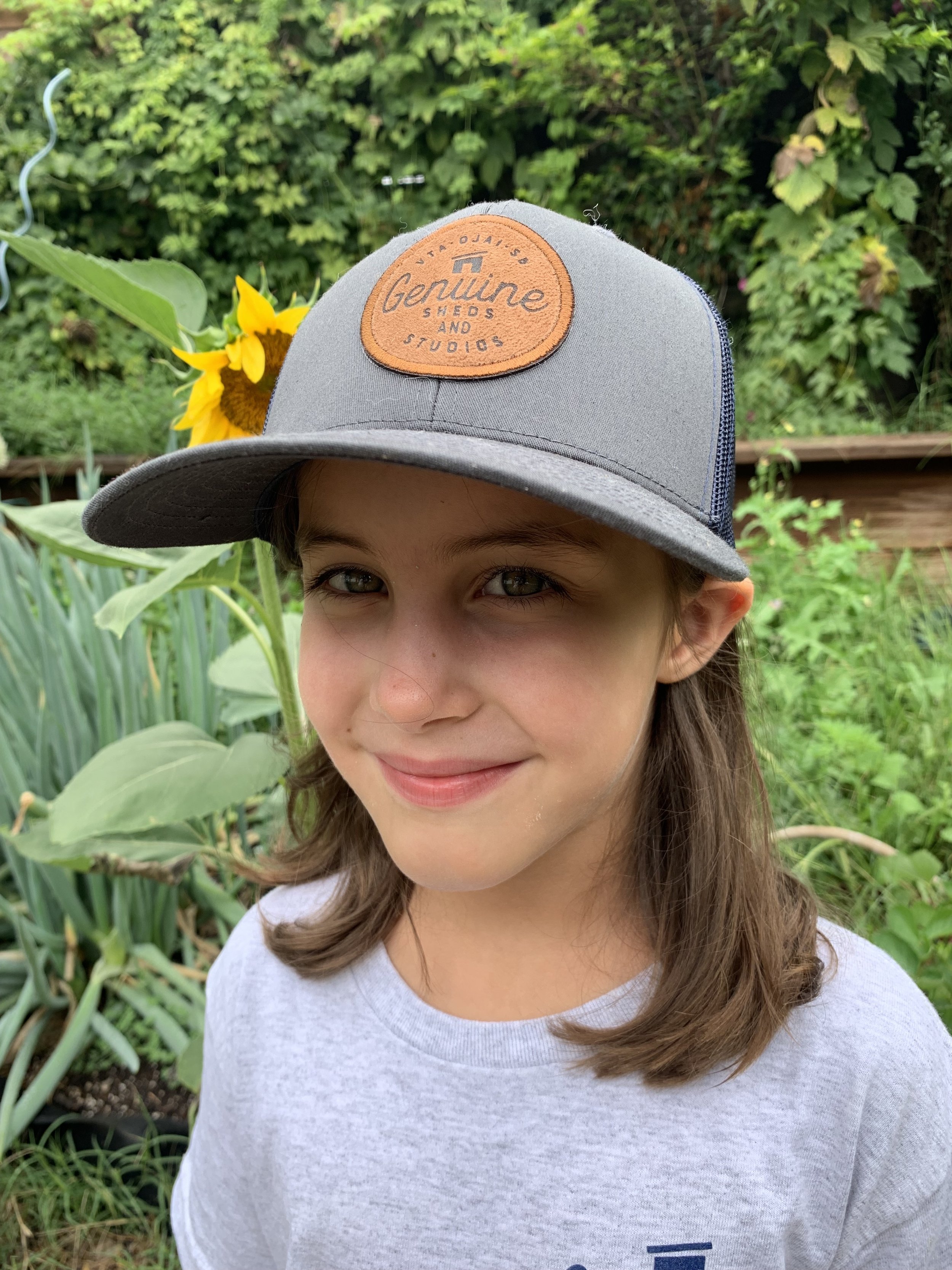 Child wearing a "Genuine Sheds and Studios" cap with a sunflower in the background, standing in a garden.