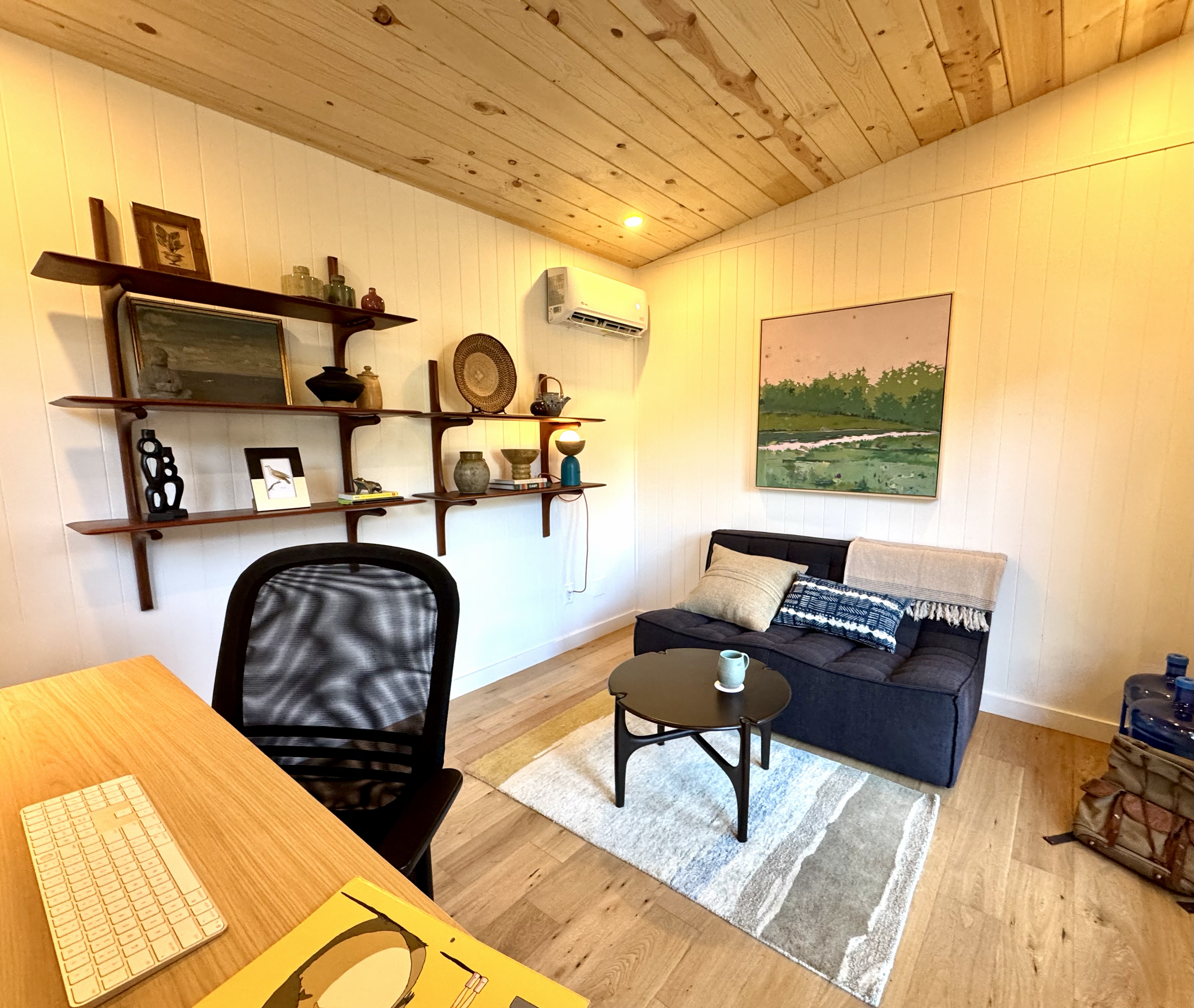 A cozy studio shed interior with wooden ceiling and white paneled walls, decorated with various art pieces and vases on wall-mounted shelves, a dark sofa with pillows, a small round coffee table. 