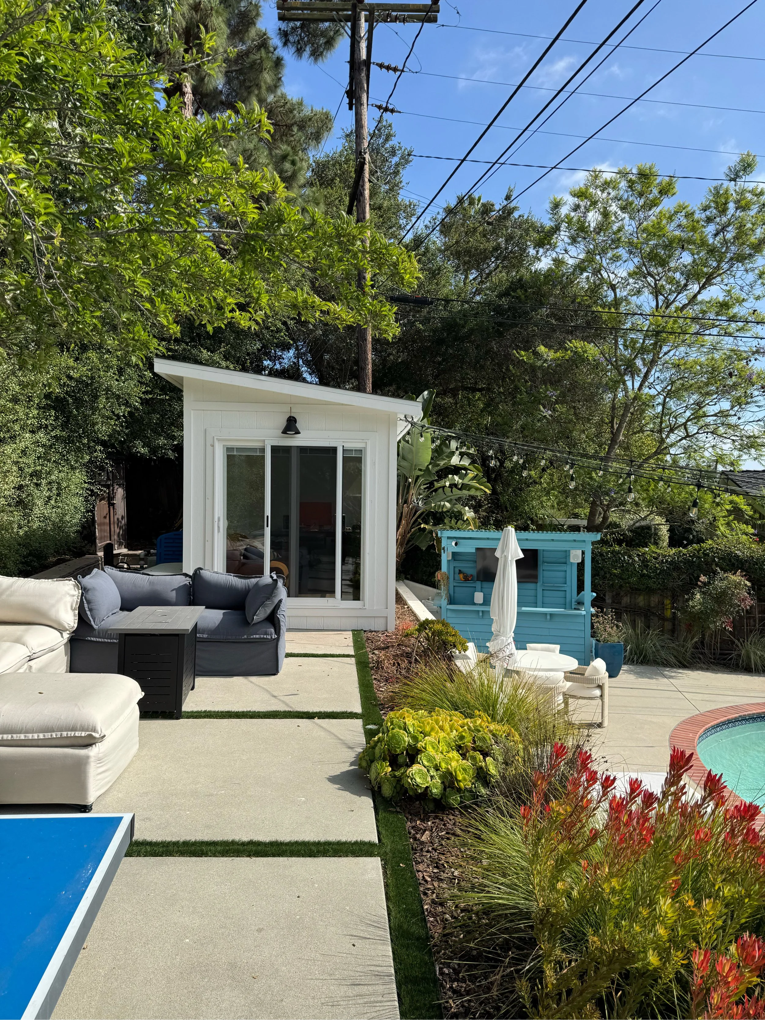 A backyard patio with outdoor seating, a small blue shed, string lights, and lush green trees and plants under a clear blue sky.