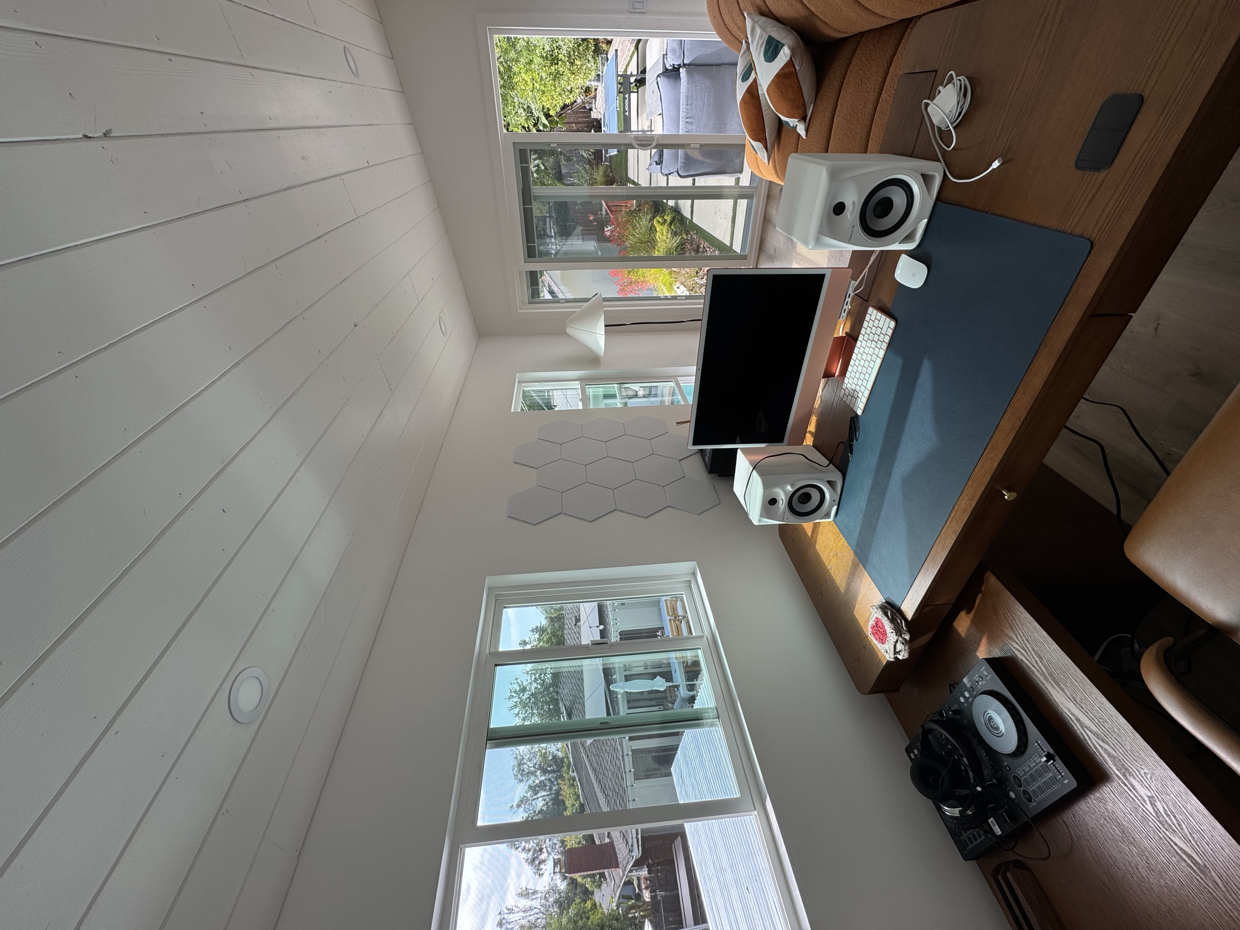 A home office with a wooden desk holding a computer, speakers, and music equipment, with large windows showing a backyard patio and garden outside.