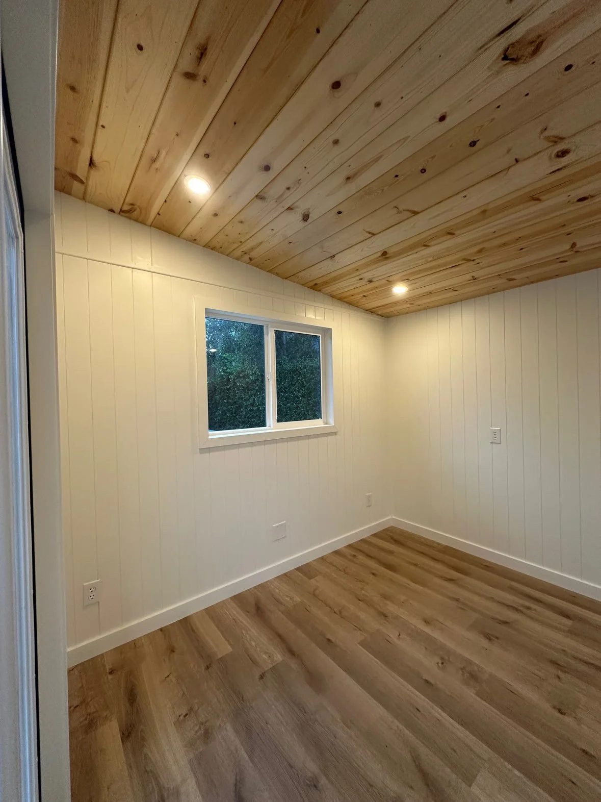 An empty room with wooden flooring, white paneled walls, a small window, and a wooden ceiling with recessed lighting.