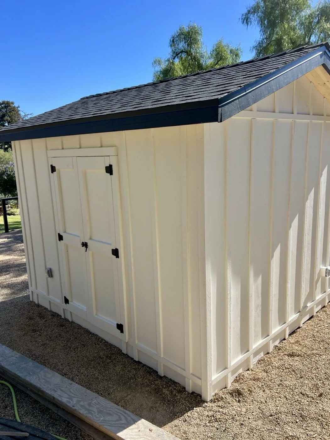 A small storage shed with cream-colored vertical siding and black hardware, situated outdoors on gravel ground with trees and blue sky in the background.