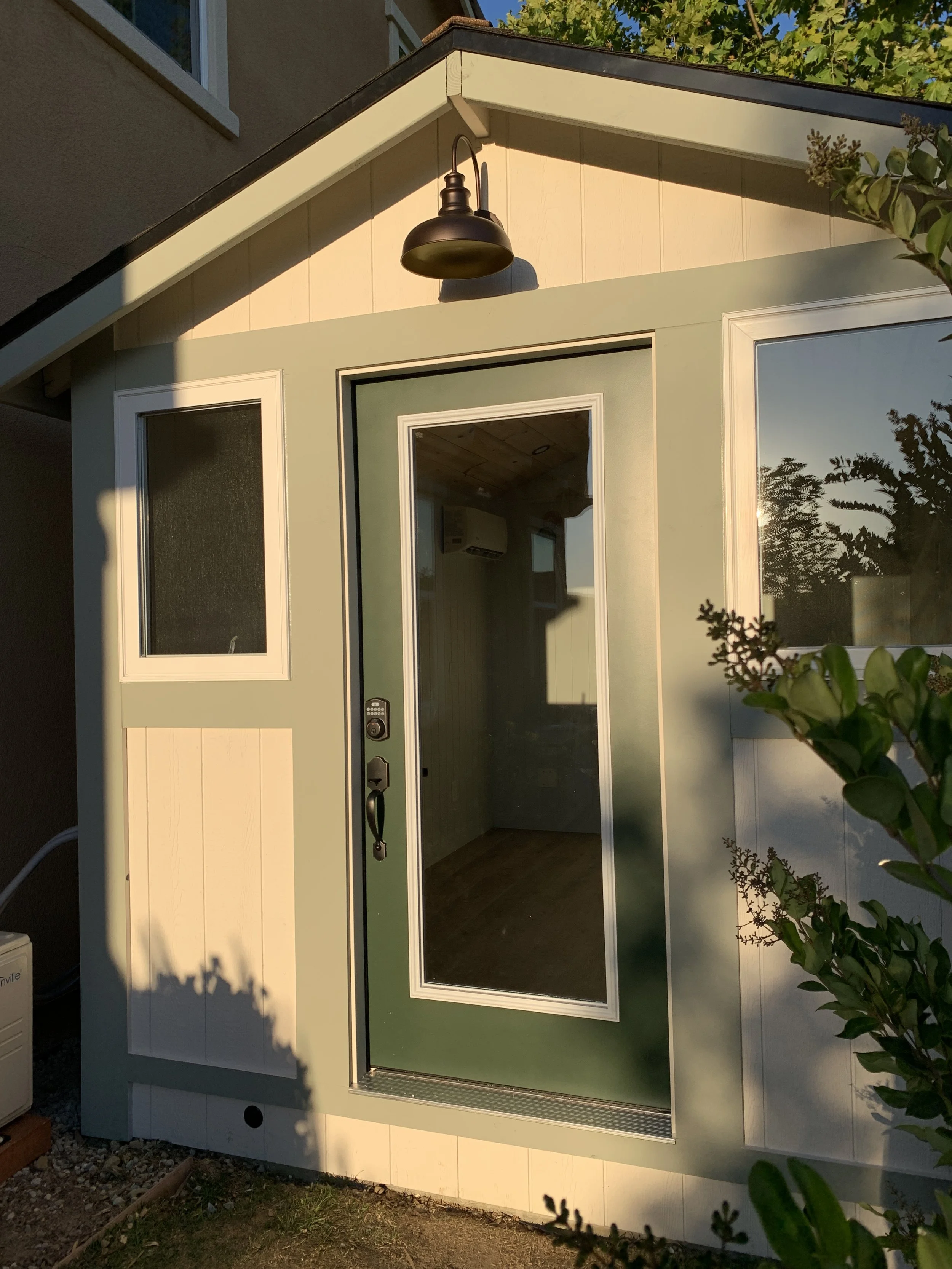 A small green shed with a large glass door, an upper window, and a black outdoor light fixture on the gable end, surrounded by garden plants.