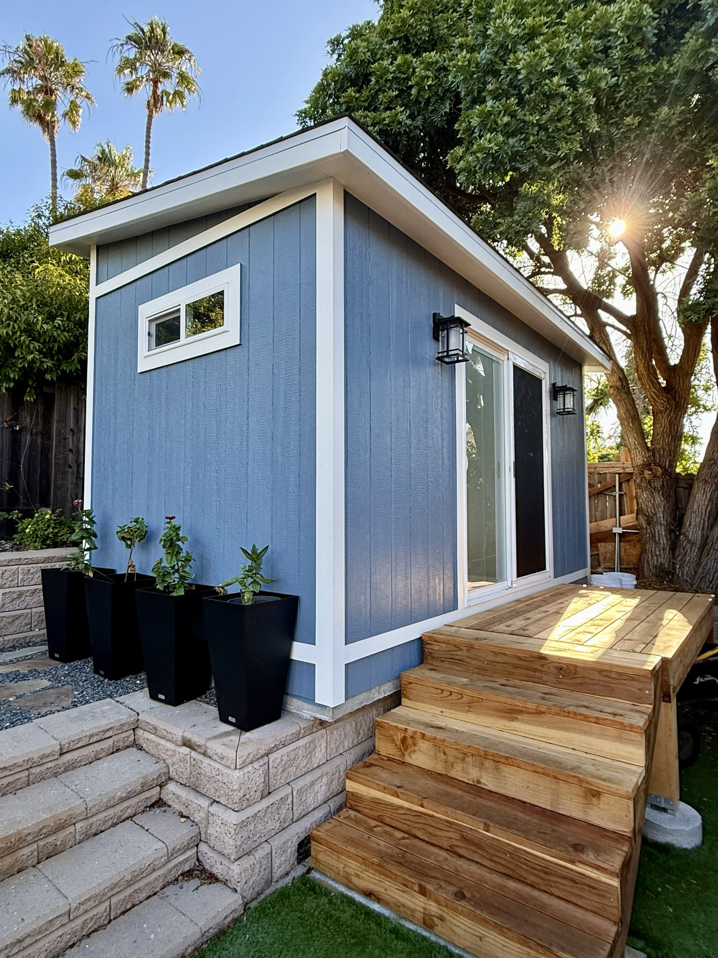 A blue studio shed with white trim and a sliding glass door, a small window near the roof, outdoor wall-mounted lanterns, a wooden deck, and wooden stairs leading up to the deck. There are trees, plants, and a fence in the background.