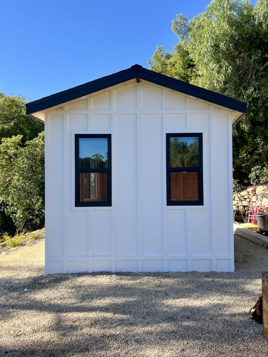 A small white wooden shed with two windows and a black roof, situated on a gravel surface with trees in the background under a clear blue sky.