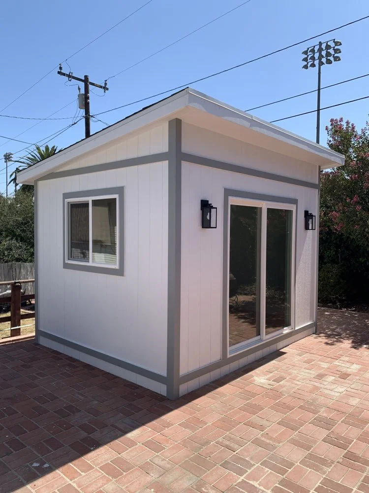 A small white shed with gray trim, a sliding glass door, a window, and two black outdoor wall-mounted lanterns, situated on a brick patio with a blue sky and some trees in the background.